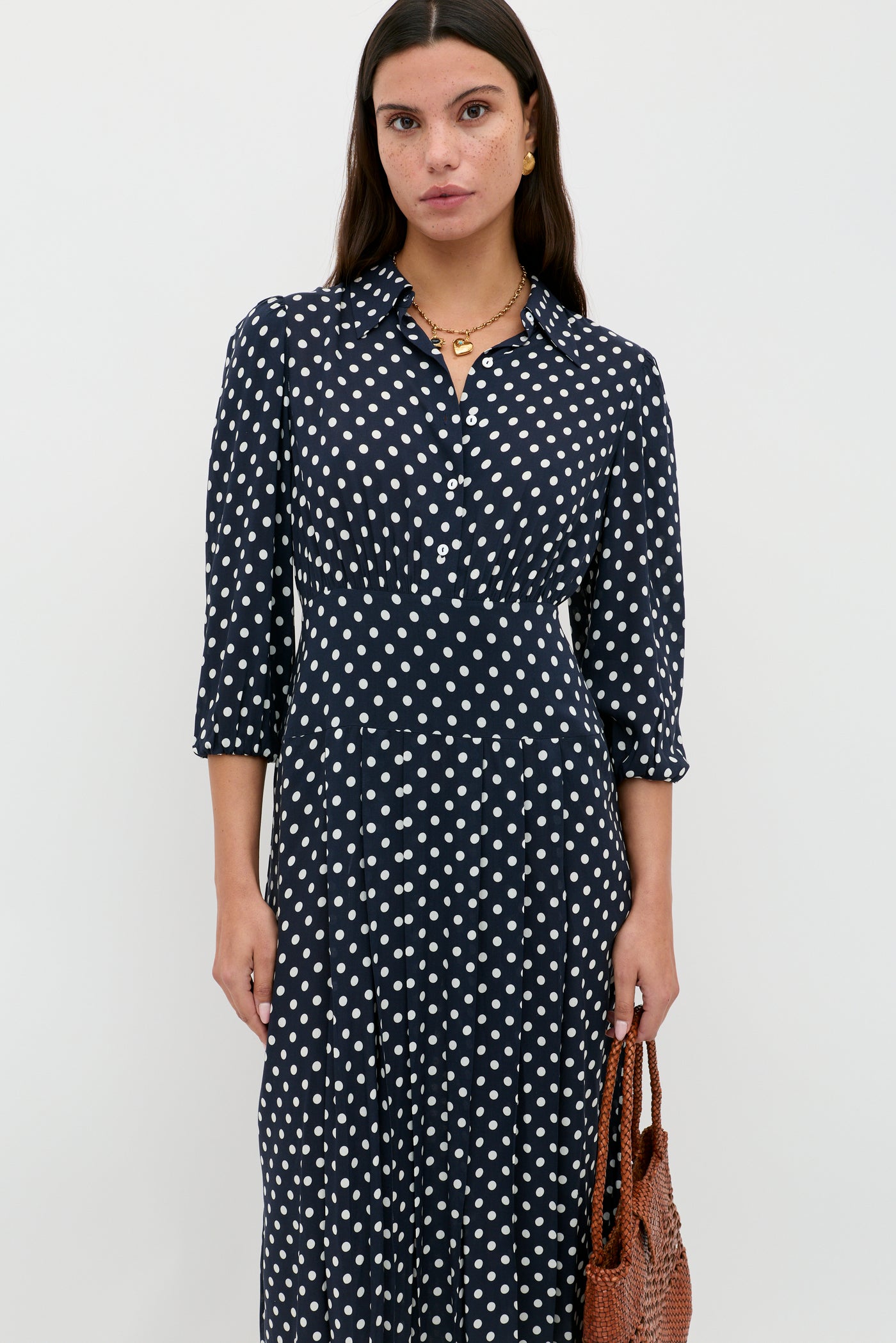 A woman stands against a plain white background wearing the Izzy - Runway Spot Navy shirtdress with blouson sleeves and a collar, holding a brown woven bag at her side.