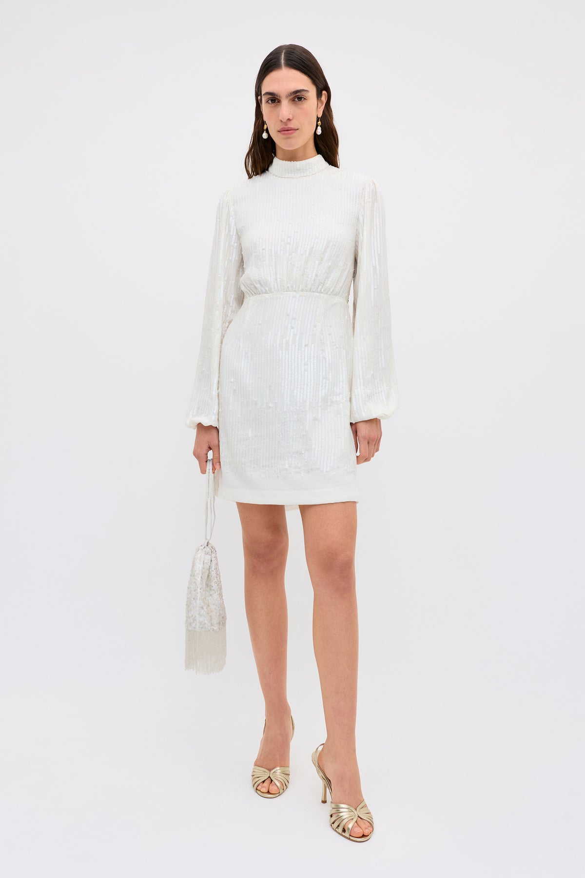 A woman wearing the Lara - Ivory Black Bow dress—long-sleeved, high-necked, white sequinned mini with black bow—holds a matching fringed purse and wears gold open-toe heels, standing against a white background.