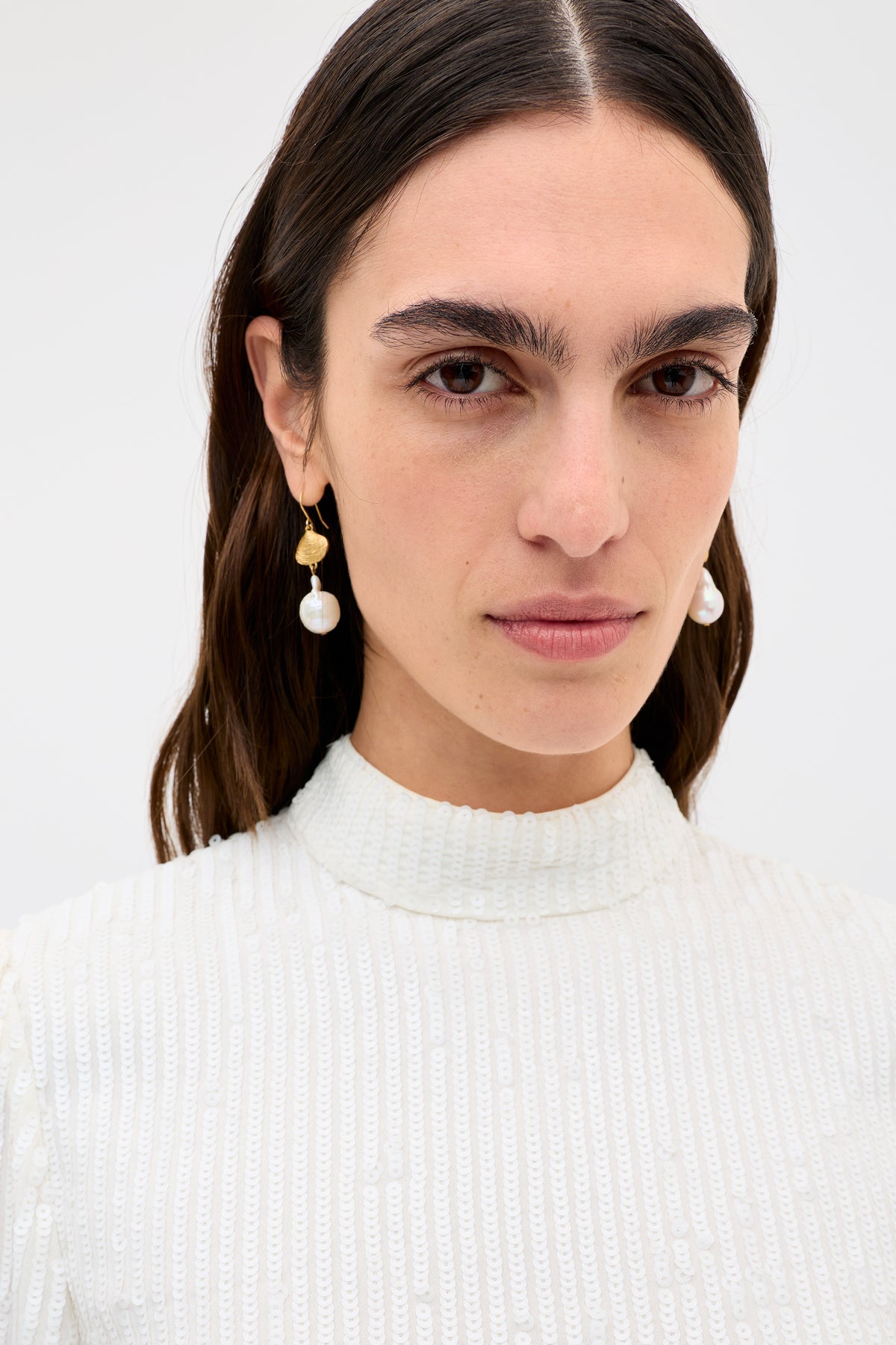 A woman with long brown hair wears the "lara - ivory black bow" high-neck textured white top and gold earrings with dangling pearls, looking directly at the camera against a plain light background.