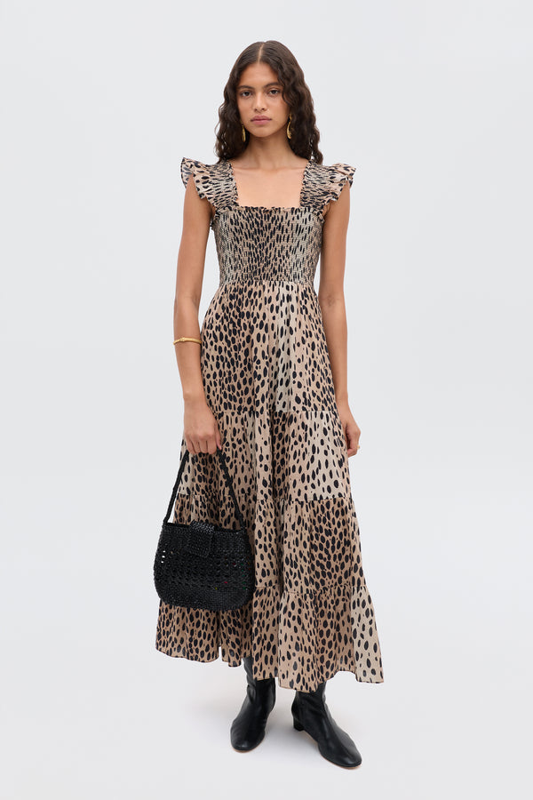A woman with long wavy hair and a neutral expression stands against a plain background wearing the Kendall - Antelope Spot Black leopard-print cotton-voile maxi dress, black ankle boots, and holding a black woven handbag.