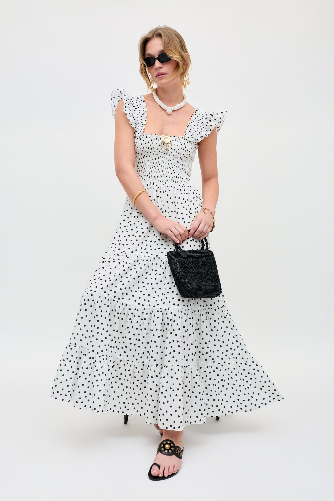 A woman wearing the Kendall - Bohemia Spot Ivory ankle-length dress with black polka dots, paired with black sandals, sunglasses, jewelry, and holding a textured black handbag stands against a plain white background.