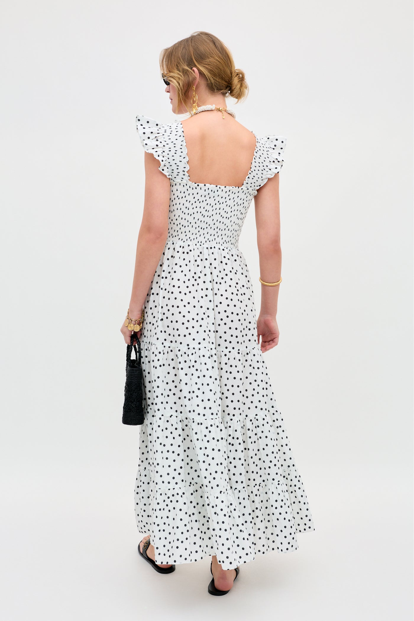 A woman with light hair in a loose updo wears the Kendall - Bohemia Spot Ivory, an ankle-length polka dot dress with ruffled sleeves and a square open back. She carries a black handbag and wears sandals against a plain light background.