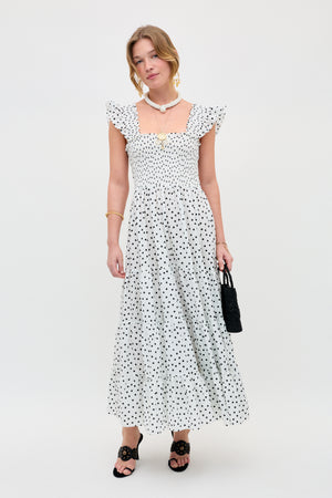 A woman wears the Kendall - Bohemia Spot Ivory dress—white with black polka dots and ruffled sleeves—paired with black sandals, a small black handbag, layered necklaces, and a bracelet, against a plain white background.