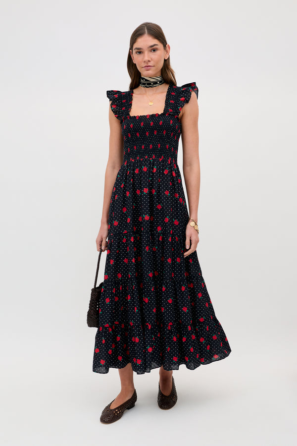 A woman stands against a plain background wearing the Kendall - Rose Spot Black, an ankle-length, sleeveless black dress with red and pink floral patterns, ruffled shoulders, a square neckline, brown flats, a neck scarf, and holding a textured handbag.