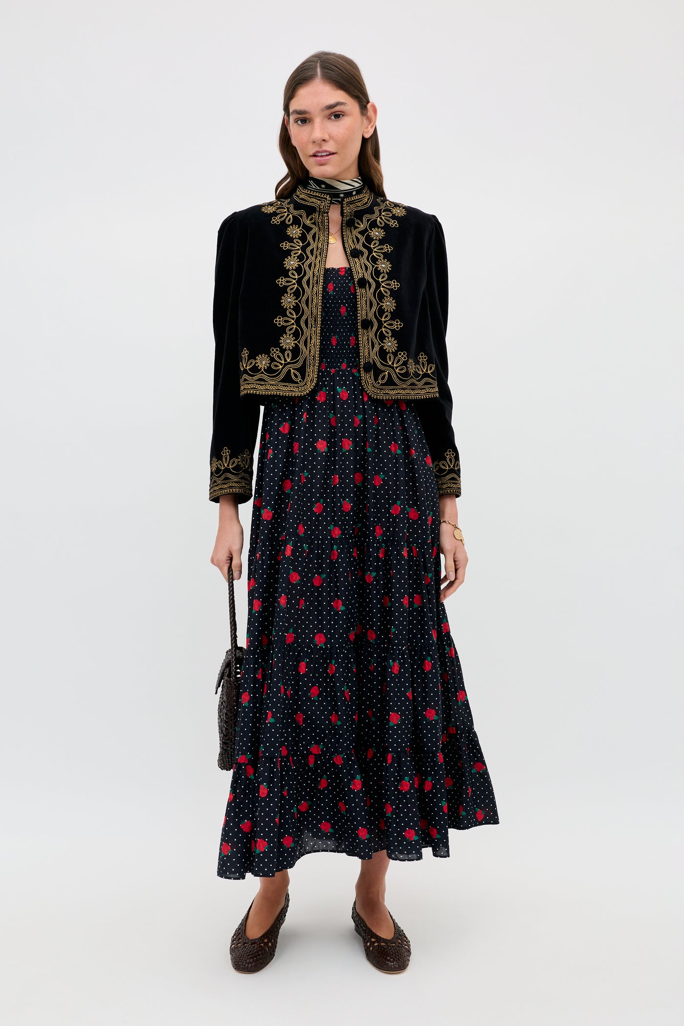 A woman stands against a plain background wearing the Kendall - Rose Spot Black, a long dark dress with a red and white polka dot pattern, paired with a black cropped jacket, brown flats, and holding a small handbag.