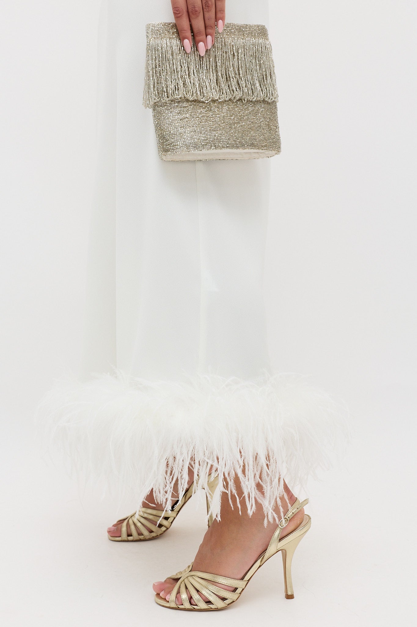 A person wearing the Mya - Ivory Feathers white skirt with ivory-feather trim, paired with gold strappy high heels and a sparkly gold fringe clutch. Only their lower body and hand are visible.