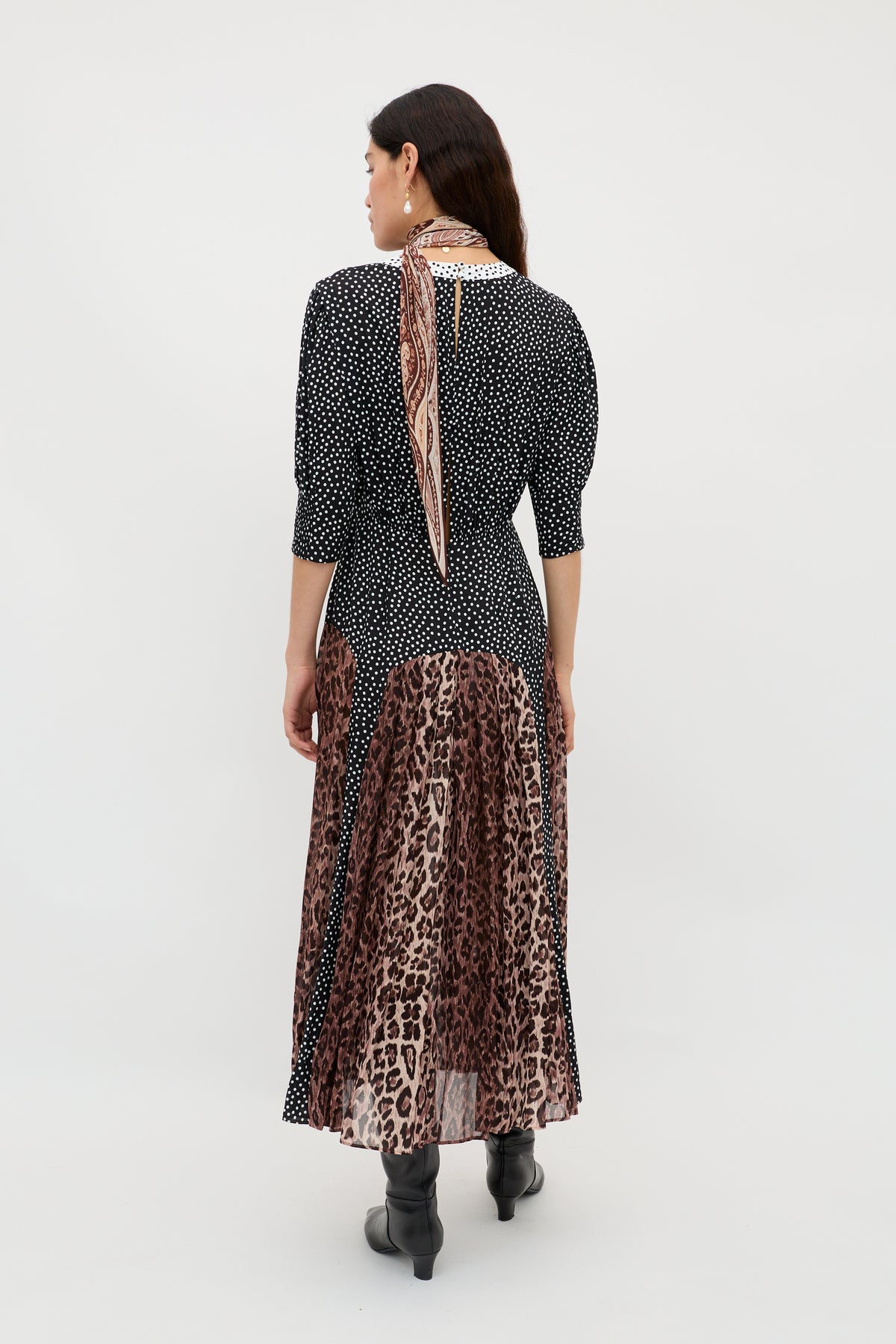 A woman faces away in the meg - leopard polka dot dress, featuring a round neck, black and white polka dot top, and leopard print midi skirt. She accessorizes with a patterned scarf, black boots, and has loose brown hair against a white background.