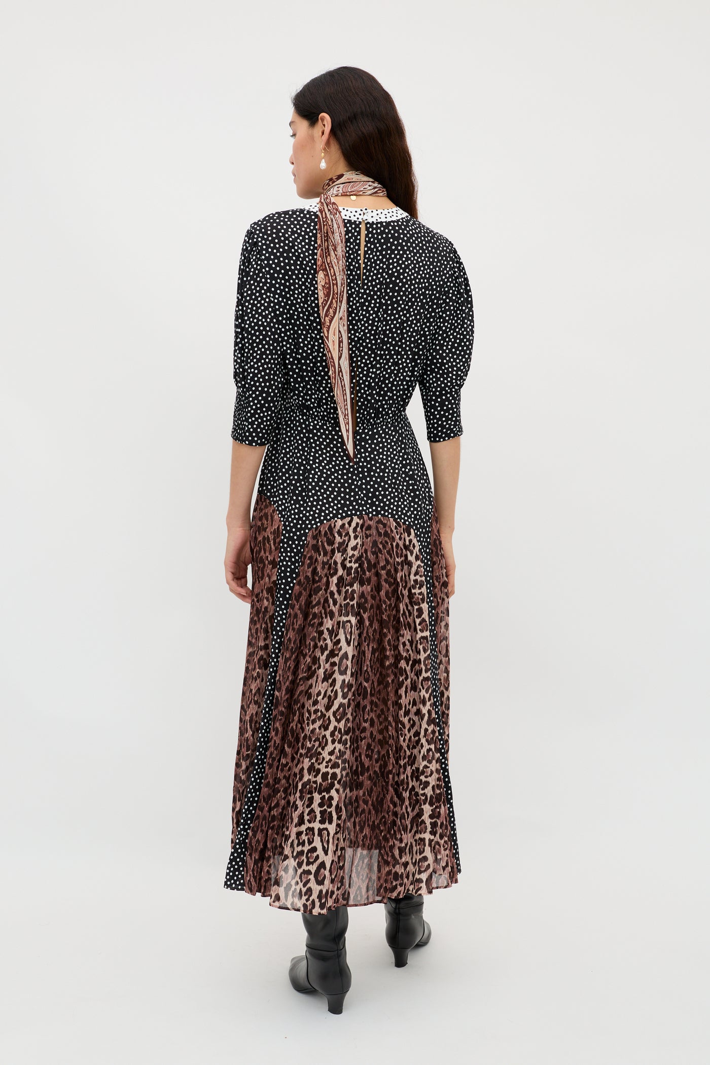A woman faces away in the meg - leopard polka dot dress, featuring a round neck, black and white polka dot top, and leopard print midi skirt. She accessorizes with a patterned scarf, black boots, and has loose brown hair against a white background.