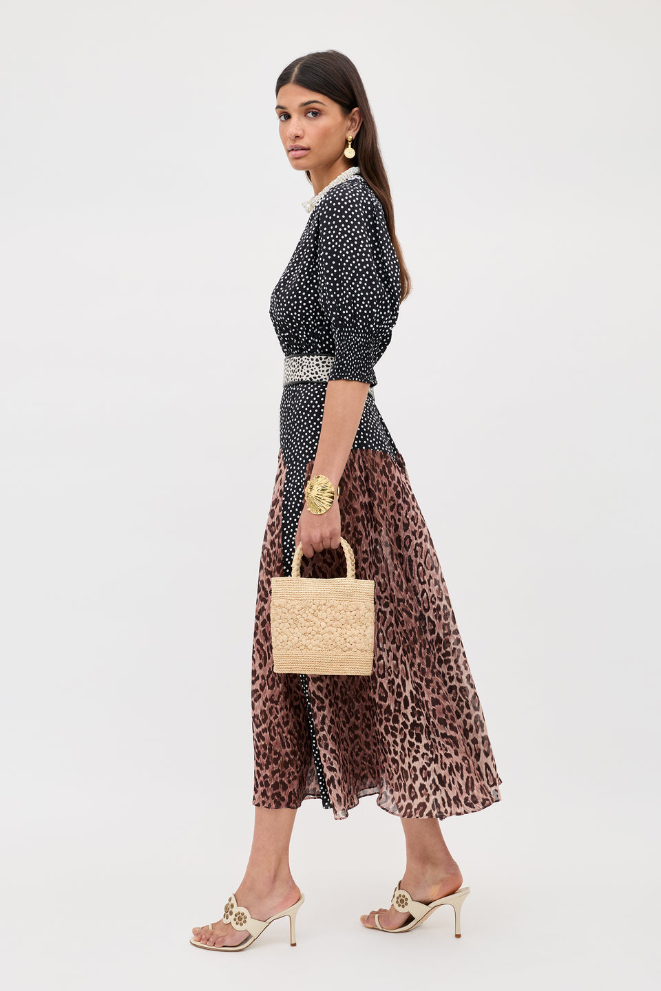 Woman in profile against a plain background, wearing the meg - leopard polka dot round-neck crepe dress, heeled sandals, gold jewelry, and carrying a woven square handbag.