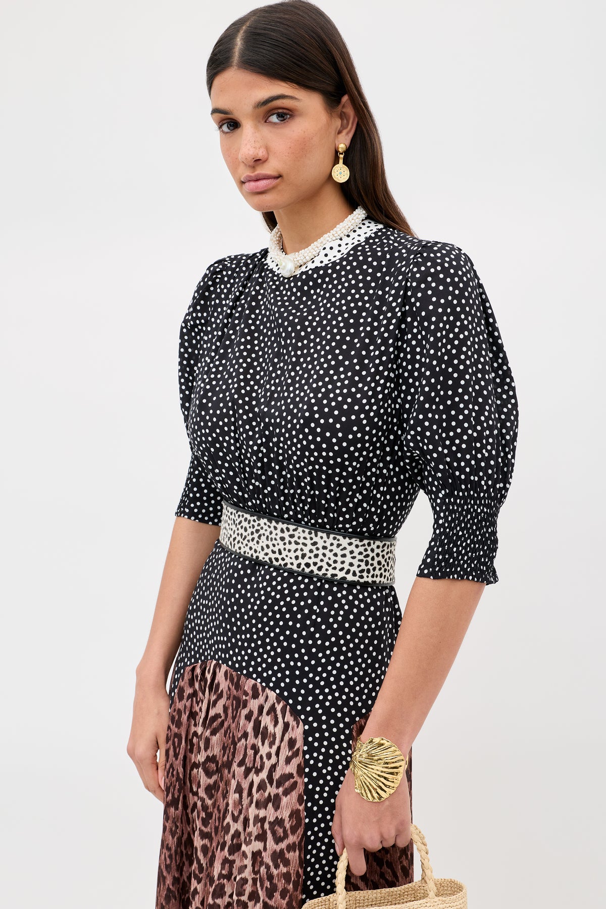 A woman with long dark hair wears the "meg - leopard polka dot" dress featuring a beaded collar, puffed sleeves, and a contrasting leopard print midi skirt. She accessorizes with gold earrings, a gold bracelet, and carries a woven bag.