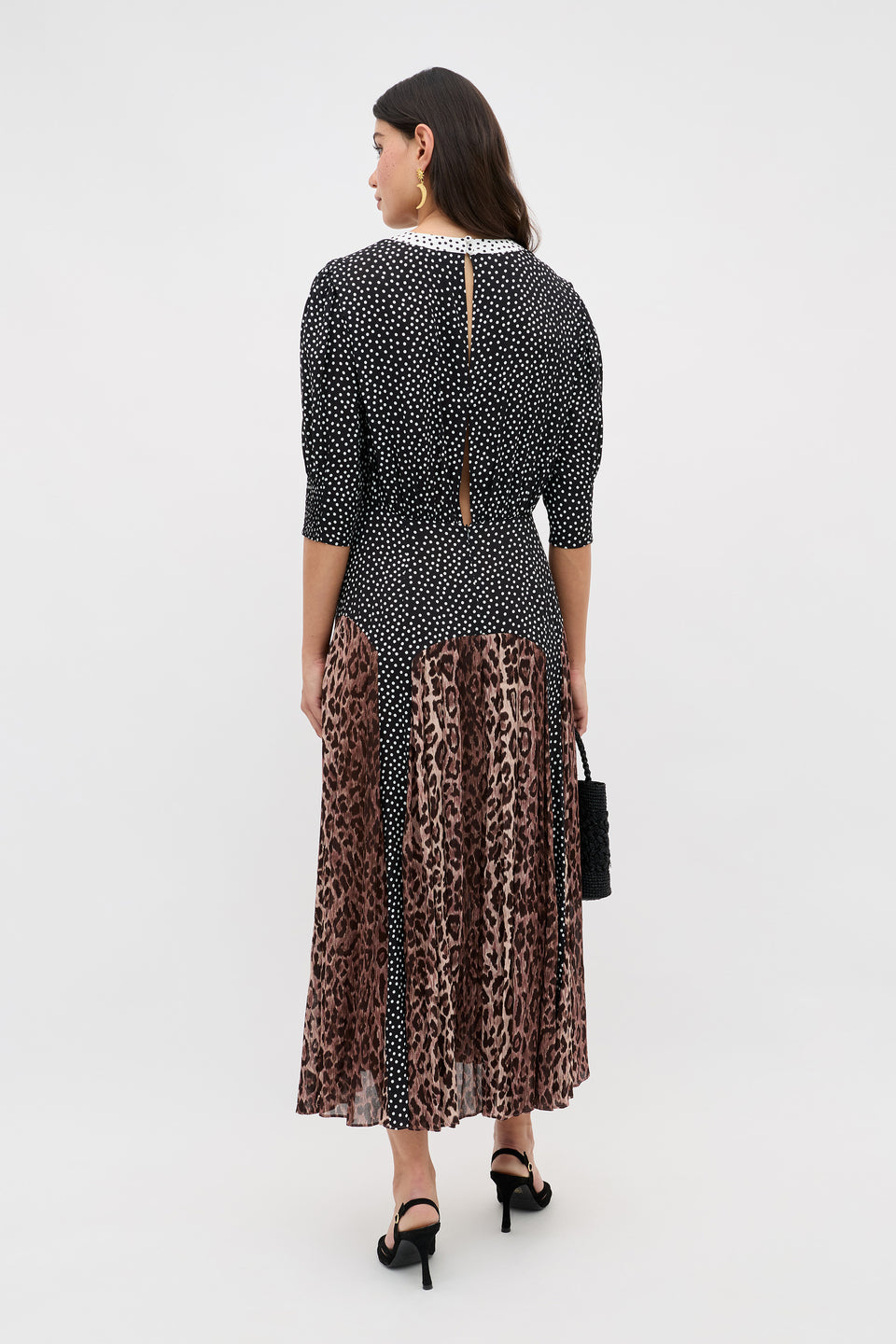 A woman faces away, wearing the meg - leopard polka dot dress featuring vintage-inspired polka dots, three-quarter sleeves, and a flowy brown leopard-print midi skirt. She carries a black bag and wears black heeled sandals.