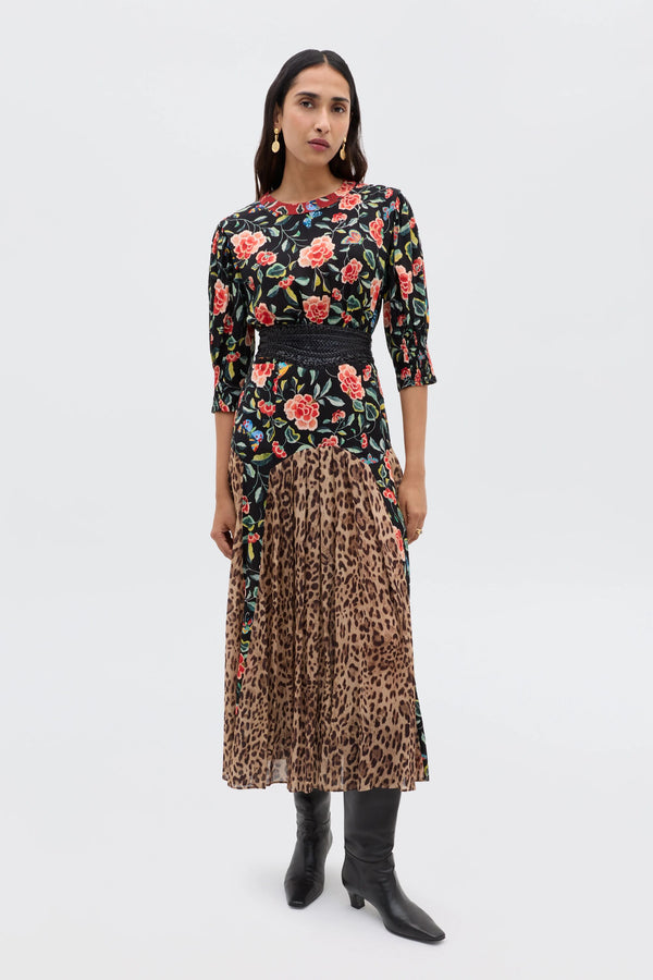 A woman stands against a plain background, embodying relaxed everyday style in the Meg - Mila Butterfly Mix Black dress by RIXO, styled with a black belt, gold earrings, and black knee-high boots.