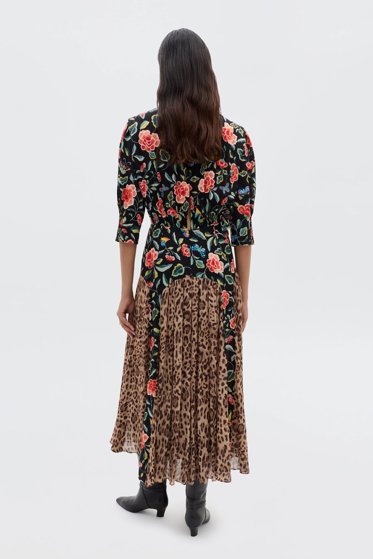 A person with long dark hair faces away, wearing the RIXO Meg - Mila Butterfly Mix Black dress, featuring a black top with red florals and a pleated leopard skirt. Black boots and a light background complete this effortlessly stylish look.
