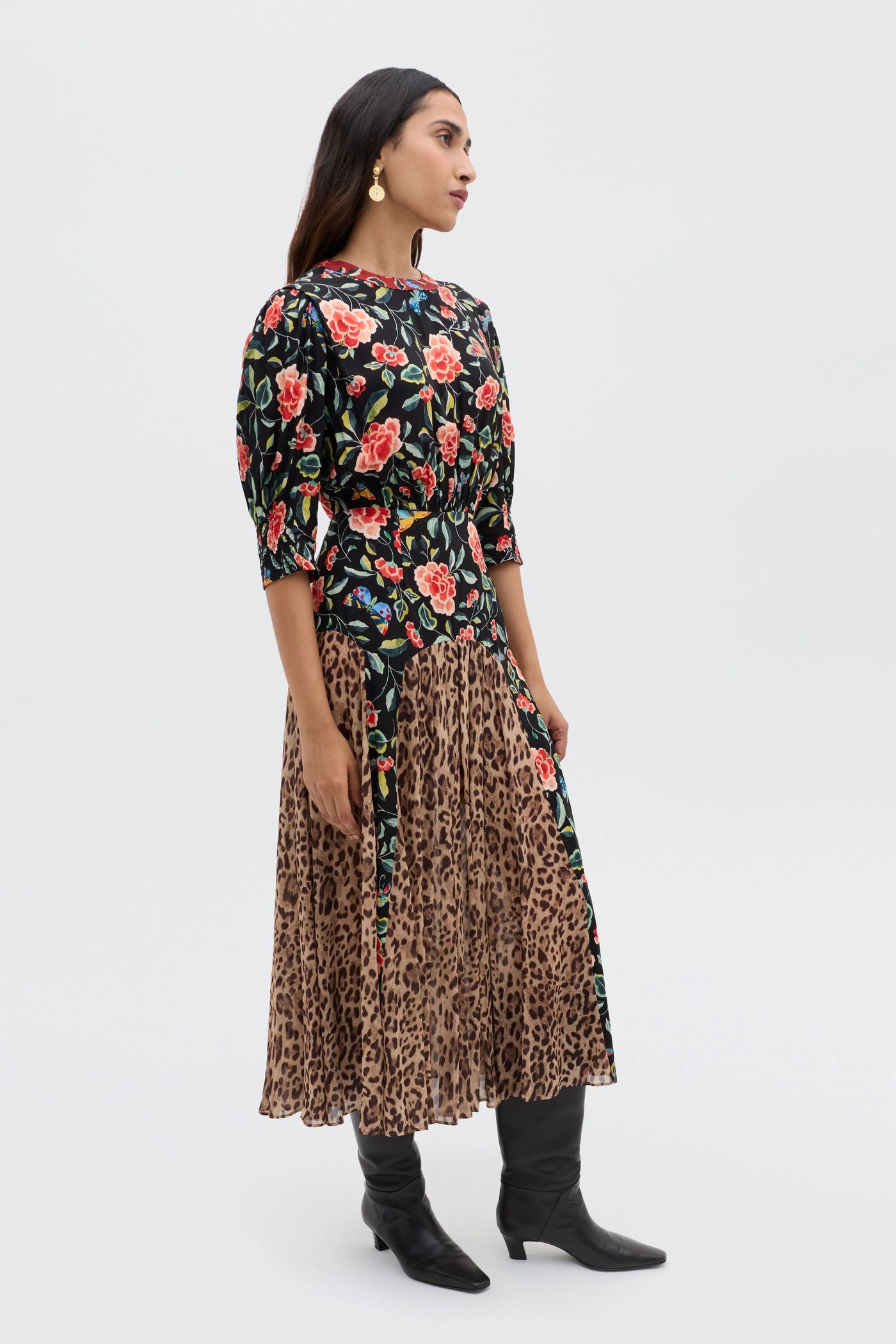 A woman stands sideways against a light background, wearing the RIXO Meg - Mila Butterfly Mix Black dress with mixed patterns, styled with black boots and gold earrings for a relaxed everyday look.