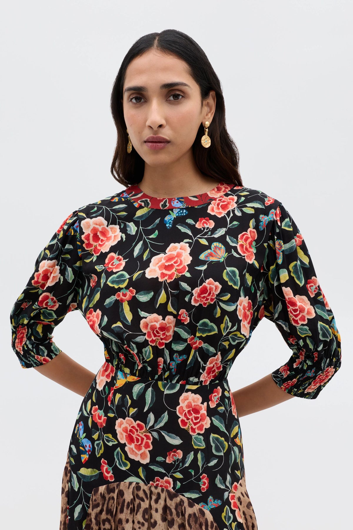 A woman with long dark hair wears gold earrings and the RIXO Meg - Mila Butterfly Mix Black dress, featuring red and pink florals, posing confidently against a plain light background for effortless everyday style.