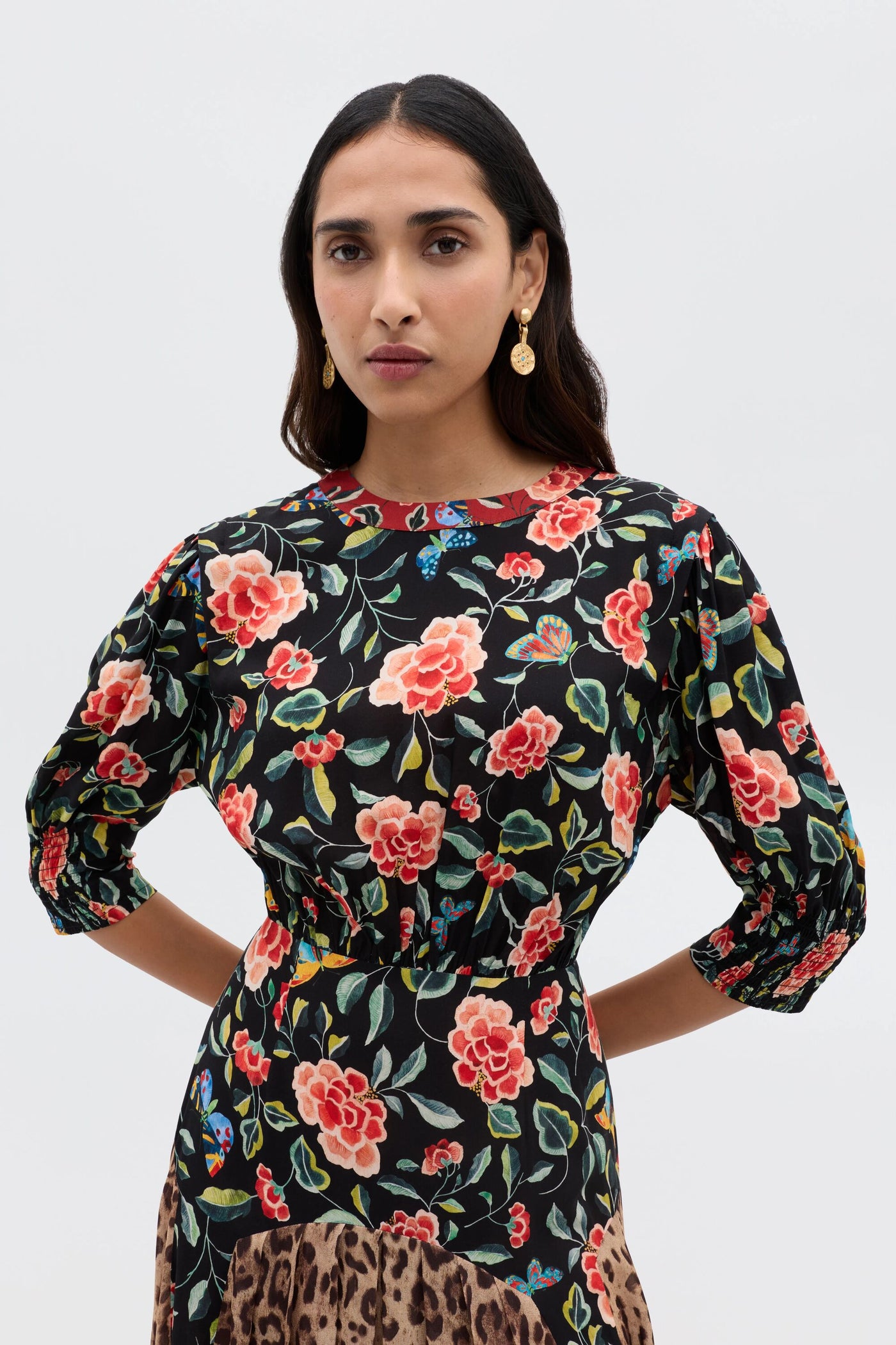 A woman with long dark hair wears gold earrings and the RIXO Meg - Mila Butterfly Mix Black dress, featuring red and pink florals, posing confidently against a plain light background for effortless everyday style.