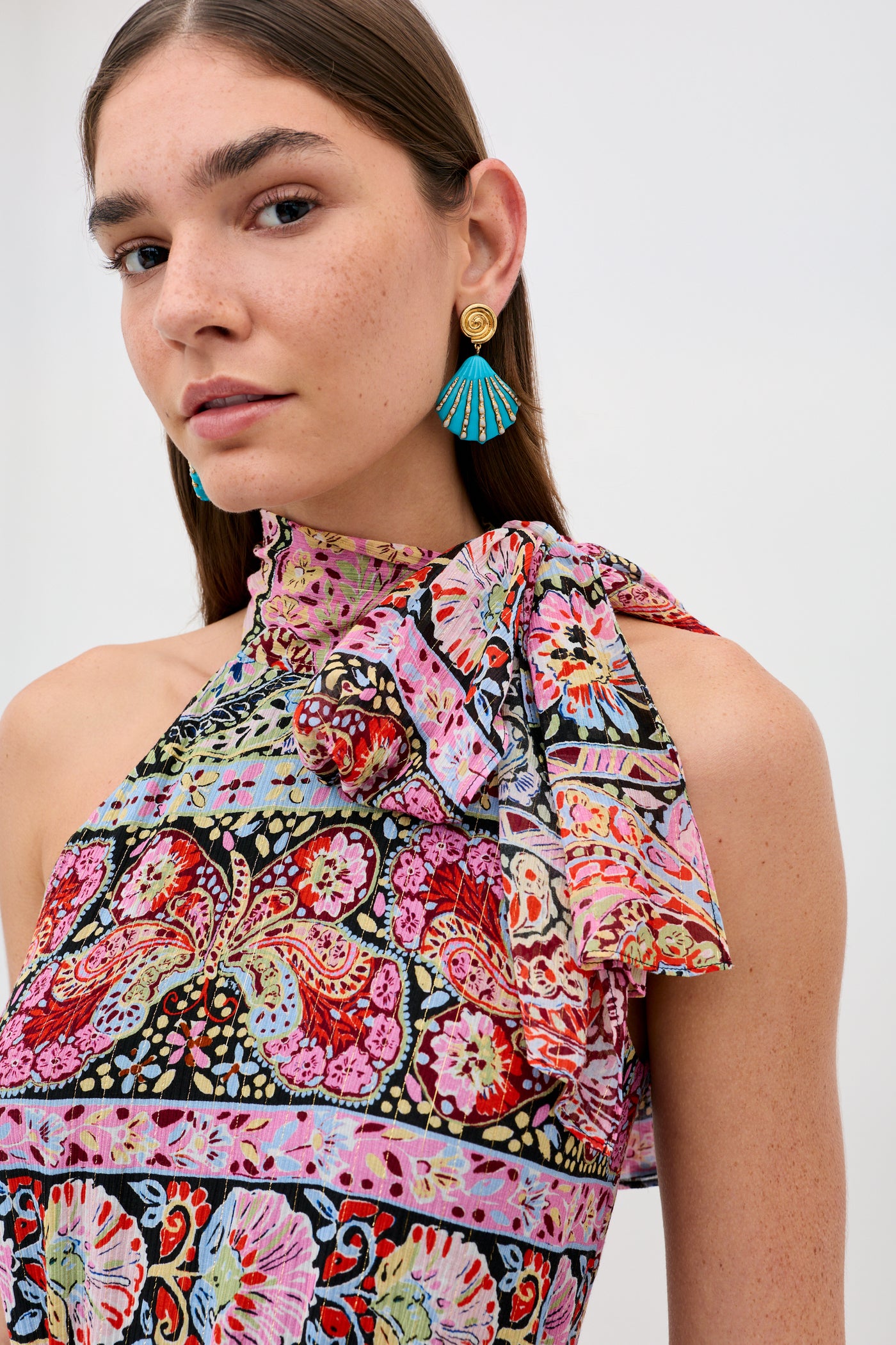 A woman with straight brown hair wears a colorful, patterned halter dress with a bow and butterfly motifs. She accessorizes with bold Daphne - Turquoise fan-shaped earrings and gold studs, against a plain, light-colored background.