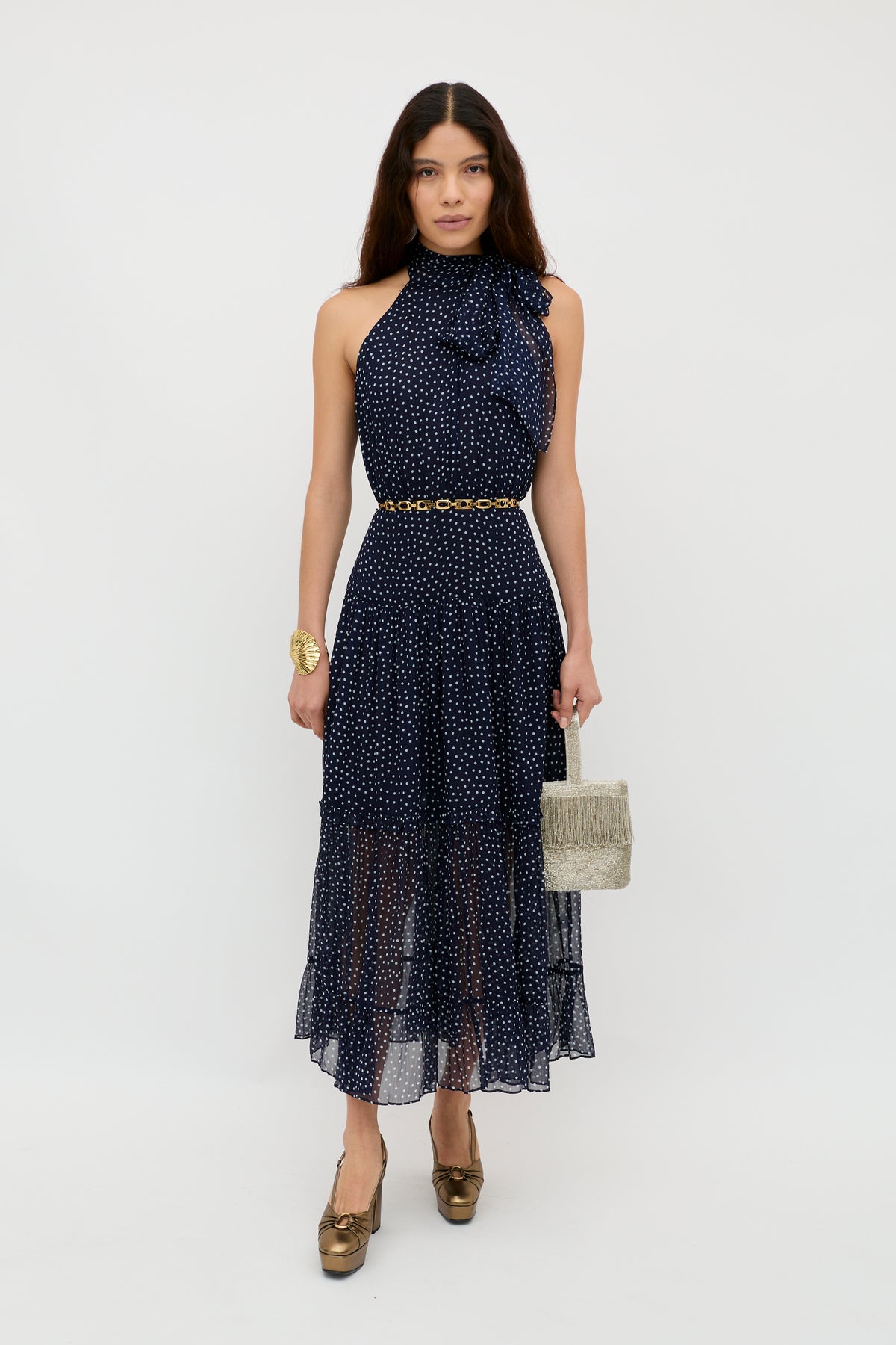 A woman poses against a white background wearing the Abi - Mini Navy Polka Dot georgette dress with halter neckline and ruffle detail, styled with a gold belt, gold heels, gold bracelet, and a textured beige handbag.