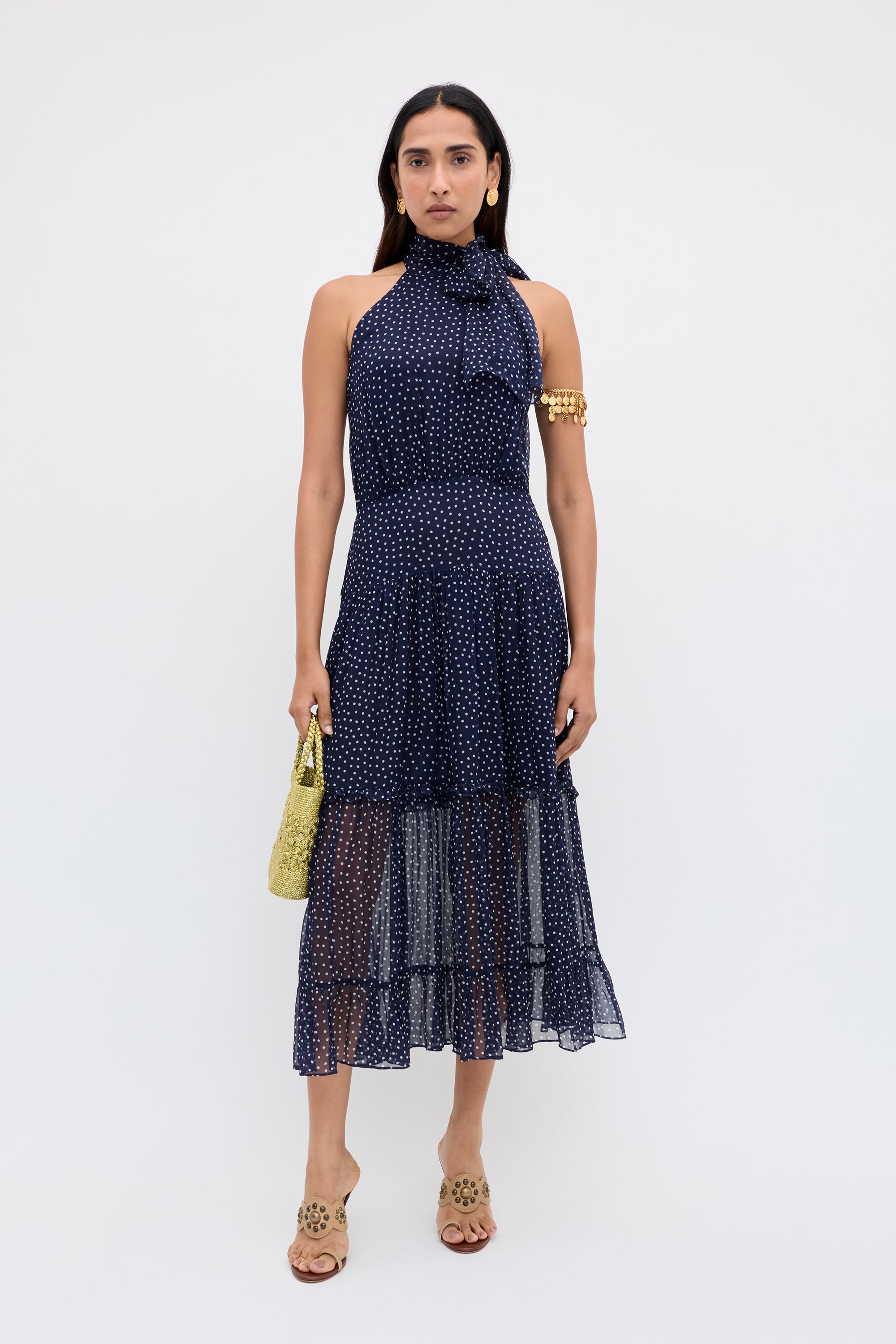 A woman stands against a white background, wearing the Abi - Mini Navy Polka Dot georgette halter-neck occasion dress with tan sandals, gold earrings, a gold arm cuff, and holding a textured yellow handbag.