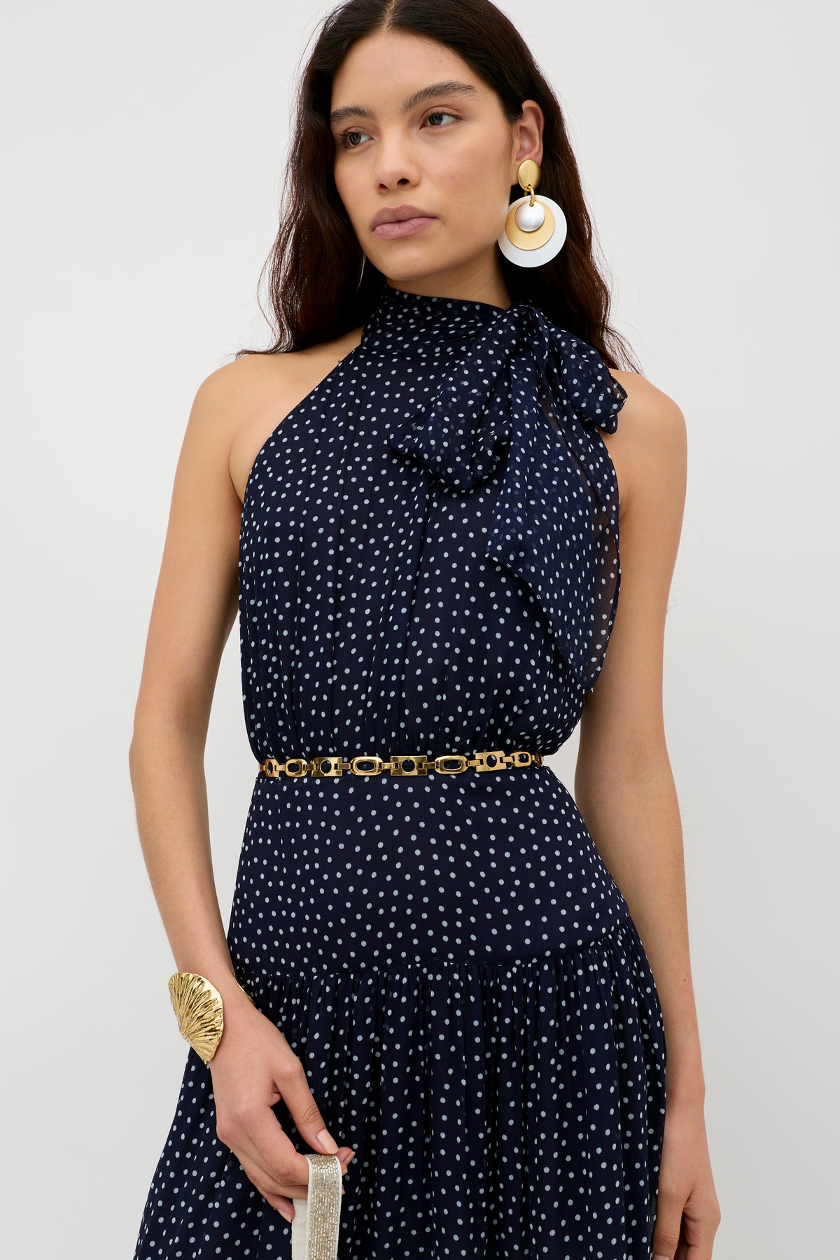 A woman wears the Abi - Mini Navy Polka Dot dress, a sleeveless georgette design with white spots, high bow neckline, gold chain belt, statement earrings, and cuff—perfect for special occasions—posed against a plain light backdrop.
