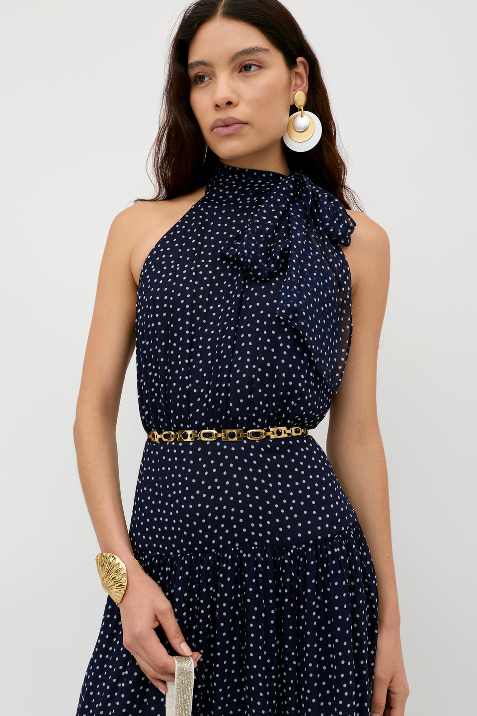 A woman wears the Abi - Mini Navy Polka Dot dress, a sleeveless georgette design with white spots, high bow neckline, gold chain belt, statement earrings, and cuff—perfect for special occasions—posed against a plain light backdrop.