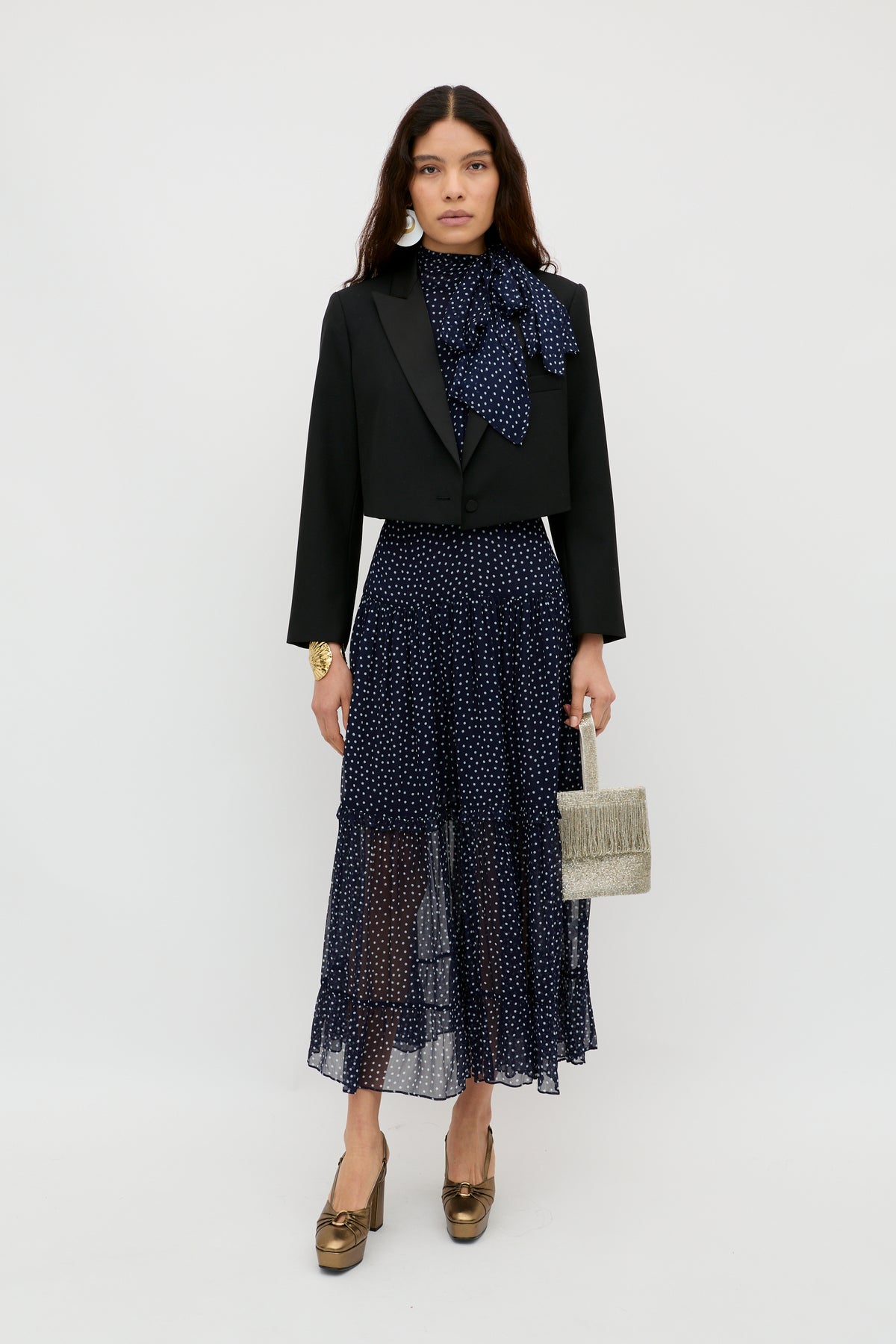 A woman stands against a plain white background wearing the Abi - Mini Navy Polka Dot dress, styled with gold heels and a small textured handbag—perfect as an occasion dress.