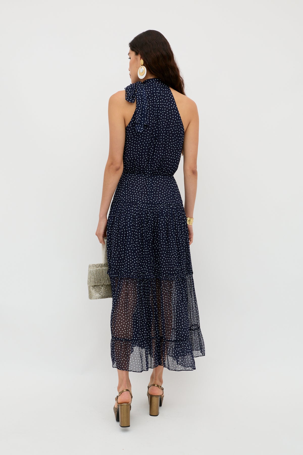 A woman with long dark hair stands facing away, wearing the Abi - Mini Navy Polka Dot dress with heeled sandals. She holds a light woven handbag and accessorizes with large hoop earrings and a gold watch against a white background.