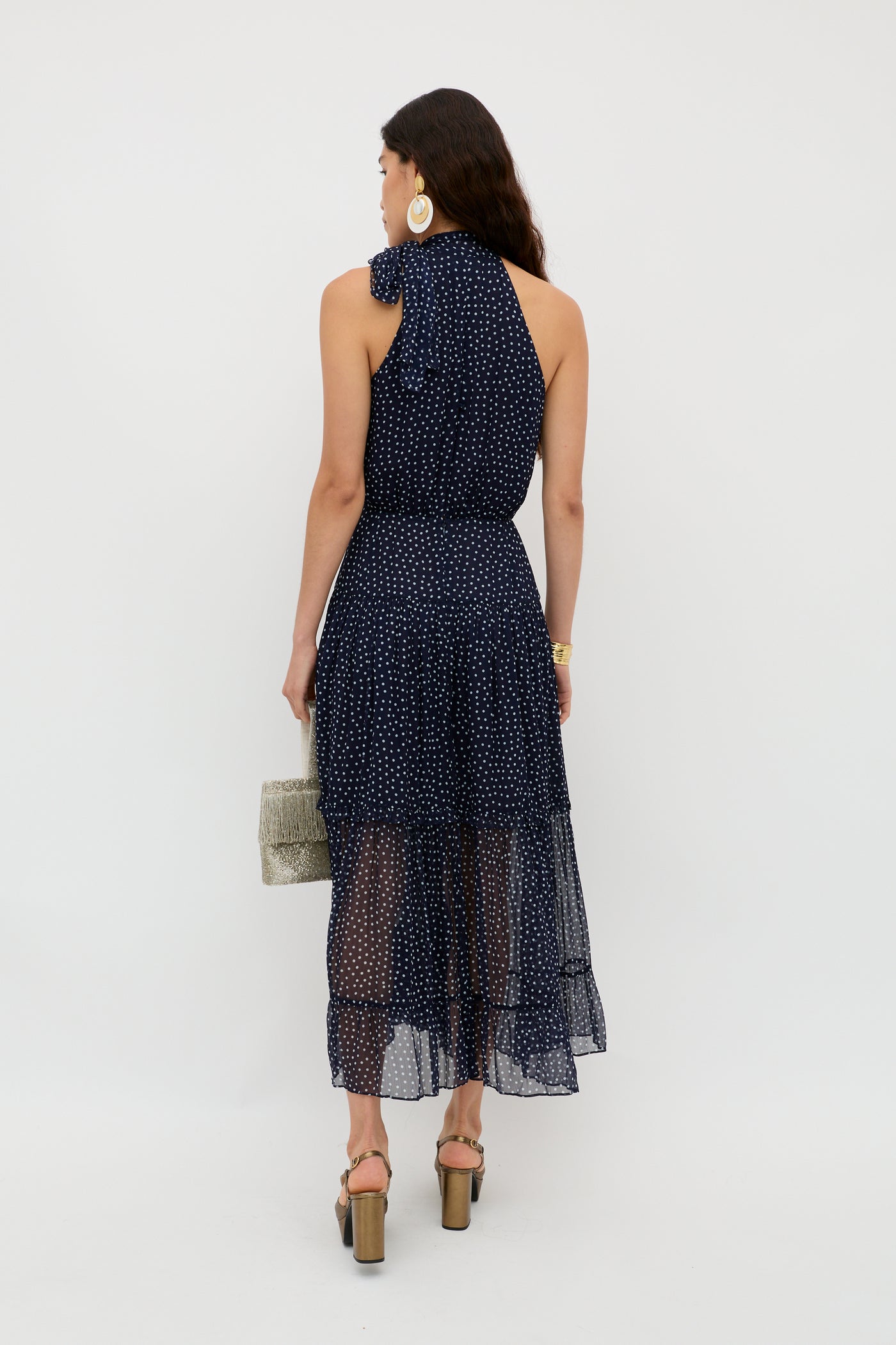 A woman with long dark hair stands facing away, wearing the Abi - Mini Navy Polka Dot dress with heeled sandals. She holds a light woven handbag and accessorizes with large hoop earrings and a gold watch against a white background.