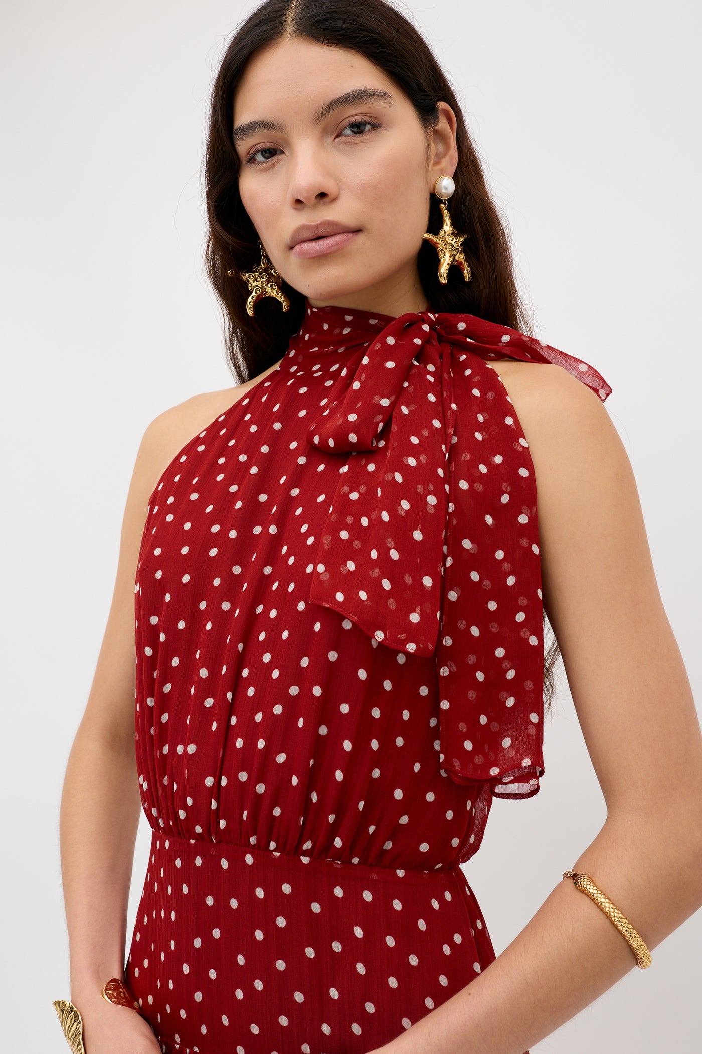 A woman with long dark hair wears the Abi - Layla Dot Red crinkled chiffon midi dress with white polka dots and a large neck bow, styled with gold sea creature earrings and a gold bracelet, standing against a plain white background.