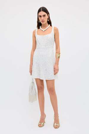A woman wears the ronan - squiggle sequin ivory sleeveless mini dress, holding a fringed handbag and accessorizing with a chunky necklace, gold statement bracelet, and gold open-toe heels, against a plain white background.