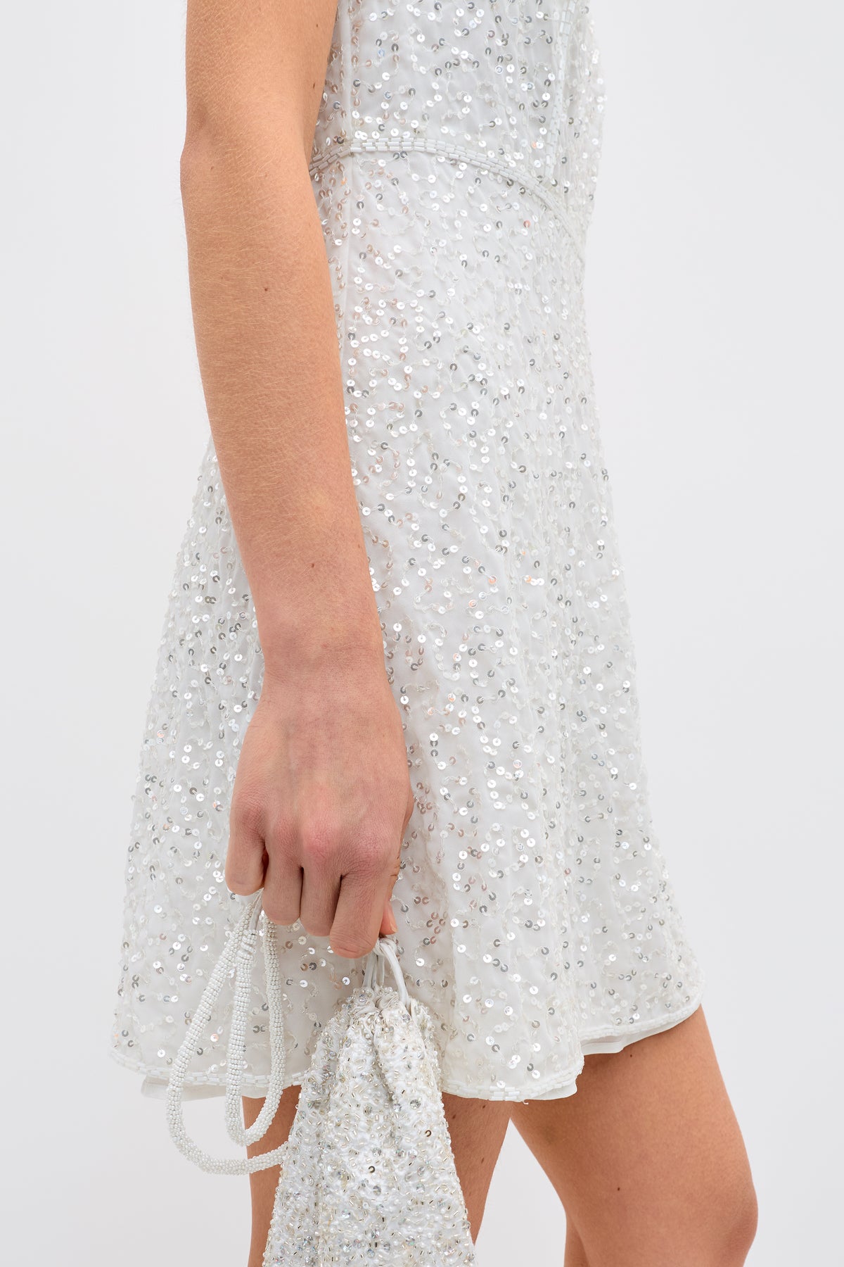 A person wears the ronan - squiggle sequin ivory sleeveless dress and holds a matching sequined drawstring bag against a plain white background.
