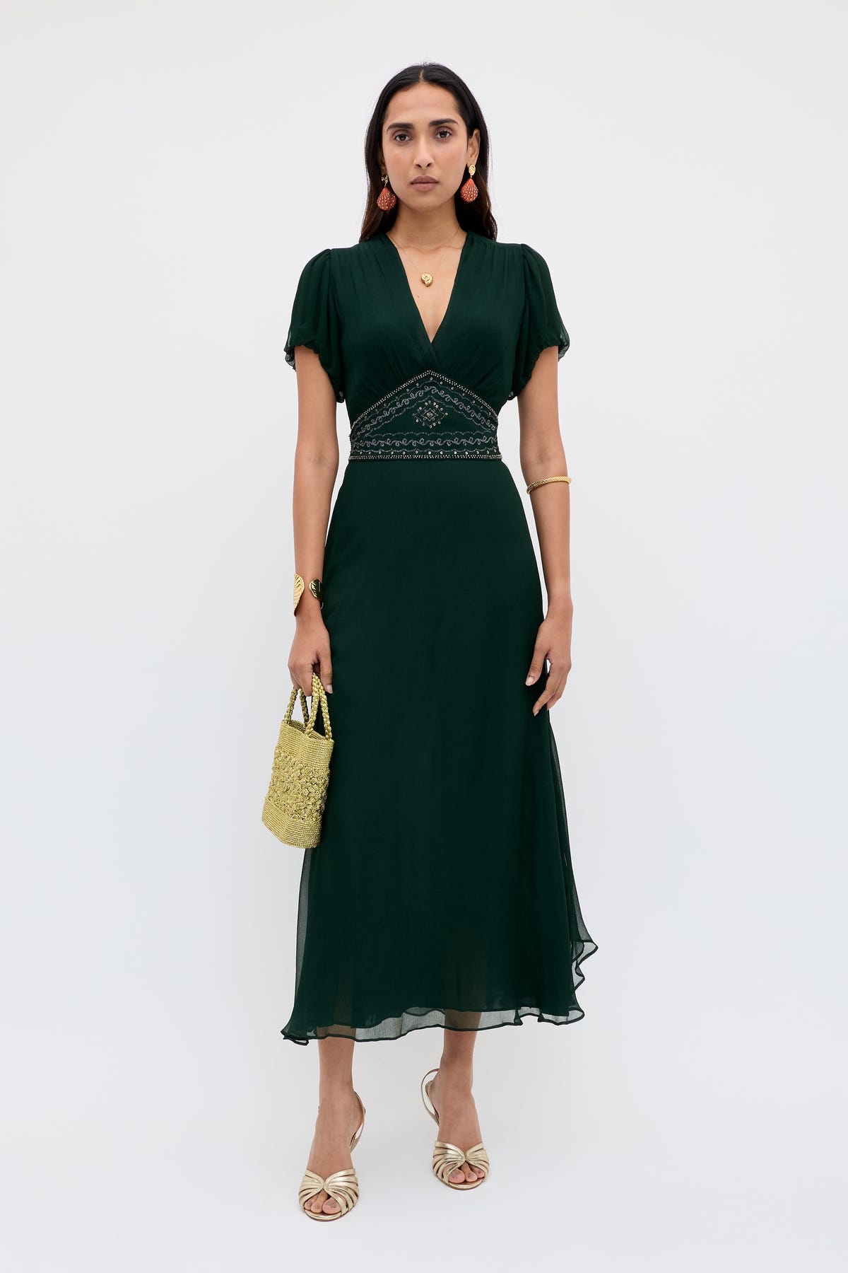 A woman poses against a white background in the Corrine - Diana Embellishment Forest Green dress, a dark green v-neck midi with embroidered waist. She accessorizes with gold sandals, bangles, earrings, a necklace, and carries a small woven bag.