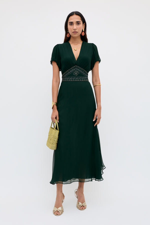 A woman poses against a white background in the Corrine - Diana Embellishment Forest Green dress, a dark green v-neck midi with embroidered waist. She accessorizes with gold sandals, bangles, earrings, a necklace, and carries a small woven bag.
