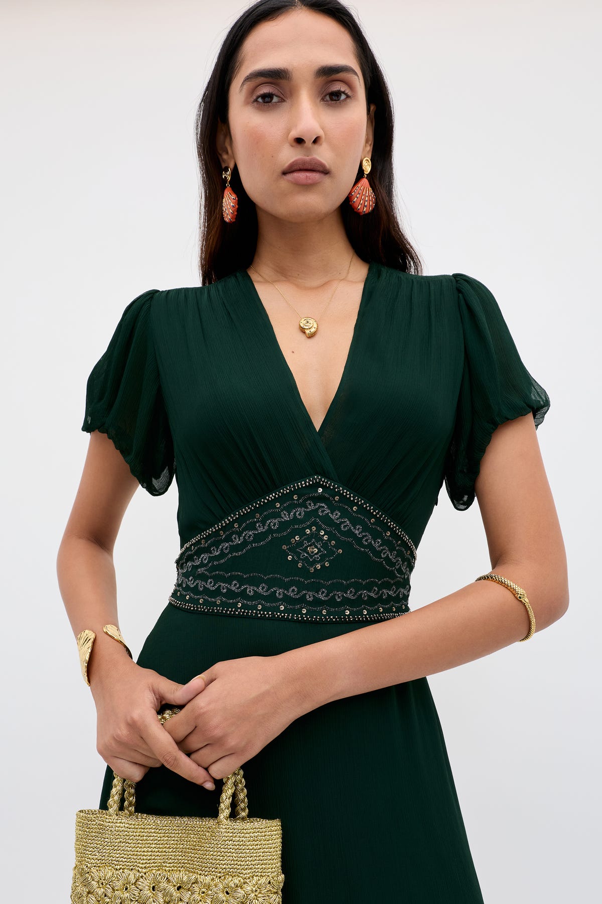 A woman wears the Corrine - Diana Embellishment Forest Green dress, featuring short sleeves, a V-neck, and a beaded waist. She accessorizes with gold jewelry, orange earrings, and a gold textured handbag against a plain light background.