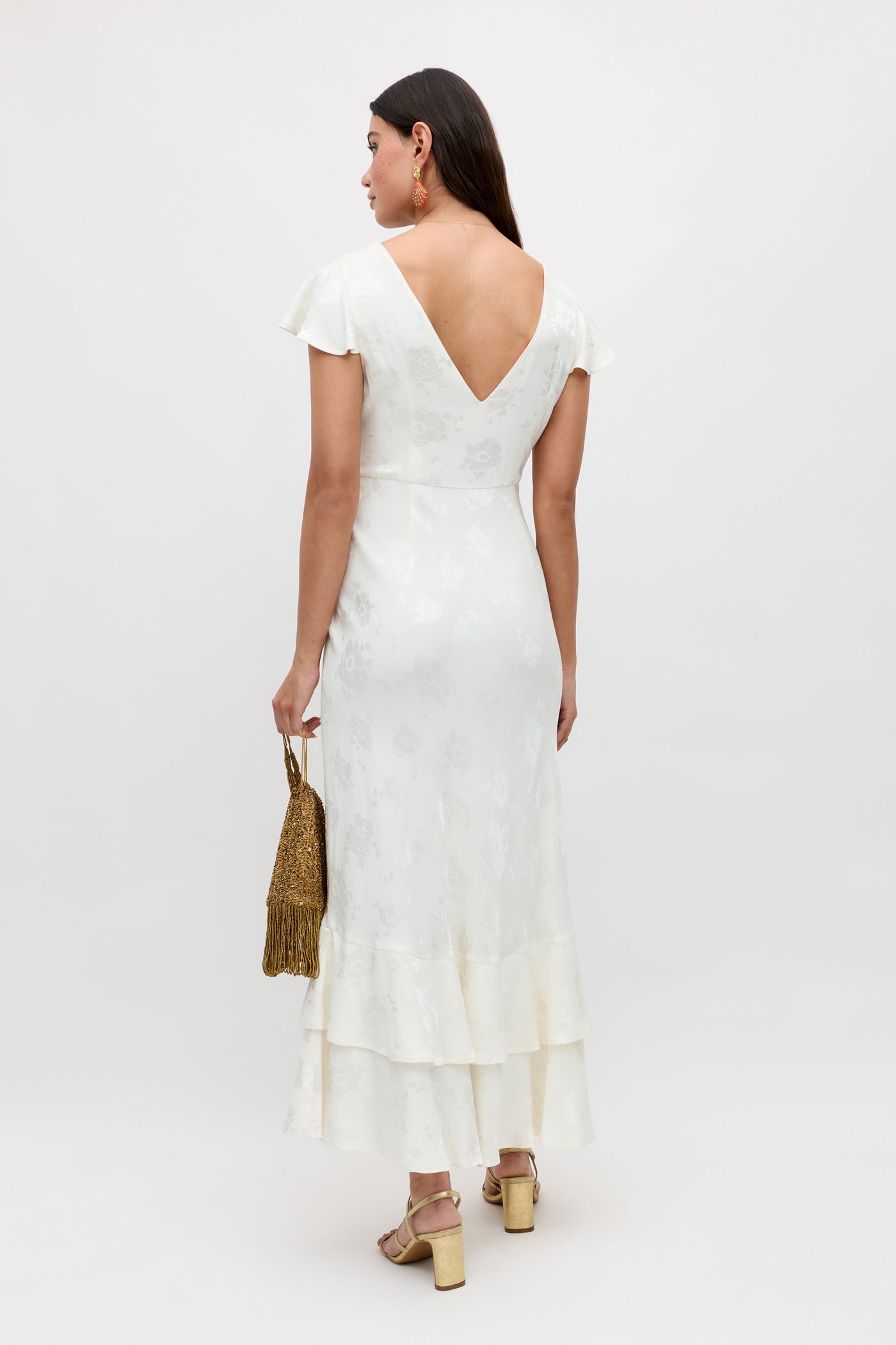 A woman faces away in the Liberty - Poppy dress, an ivory jacquard ankle-length style with cap sleeves, deep V-back, and ruffled hem. She carries a gold beaded handbag and wears heeled sandals against a plain light background.