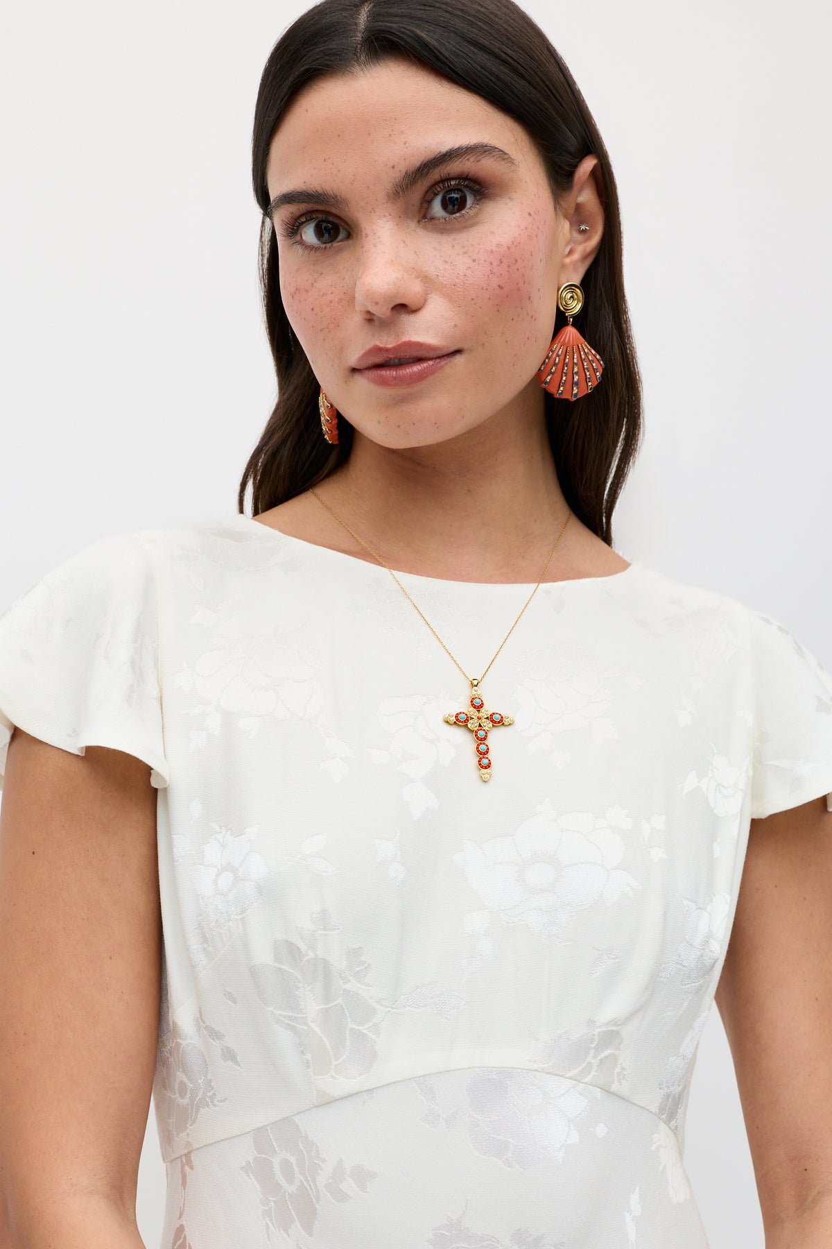 A woman wearing the Liberty - Poppy ivory jacquard floral dress with cap sleeves, gold earrings with red accents, and a gold necklace featuring a red jeweled cross pendant stands against a plain light background.