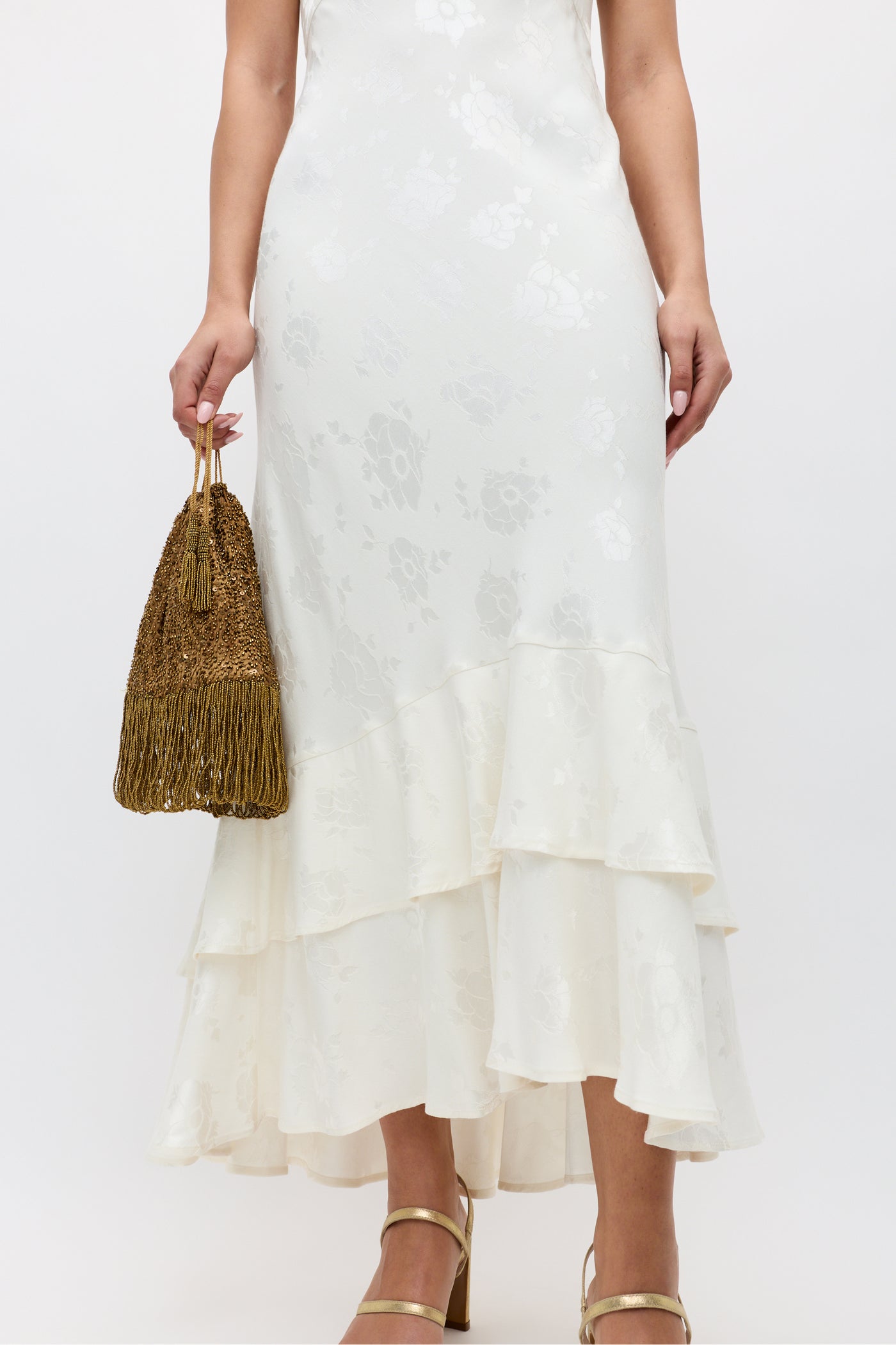 Wearing the "Liberty - Poppy" ivory jacquard floral midi dress with cap sleeves and ruffles, a woman holds a gold tasseled handbag and wears gold strappy heels. The image, cropped from shoulders to feet, showcases her elegant style.