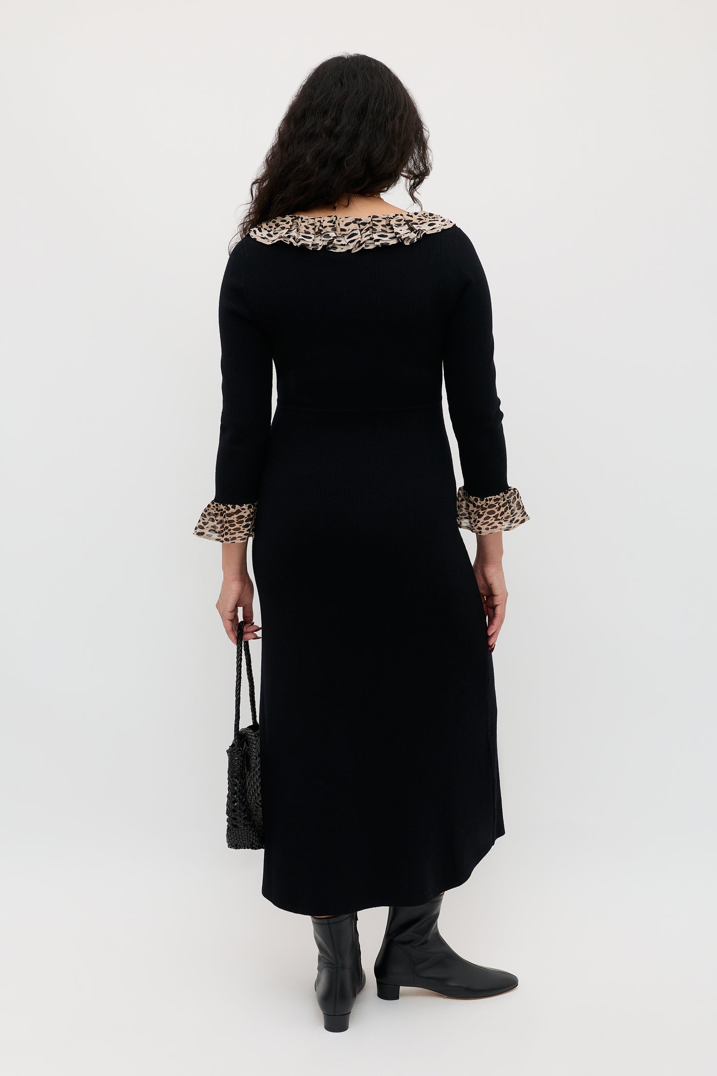 A person with long, wavy hair faces away, wearing the Mika - Antelope Spot Black merino wool dress featuring ruffled leopard trim on the collar and cuffs. They hold a black handbag and wear black boots against a plain white background.