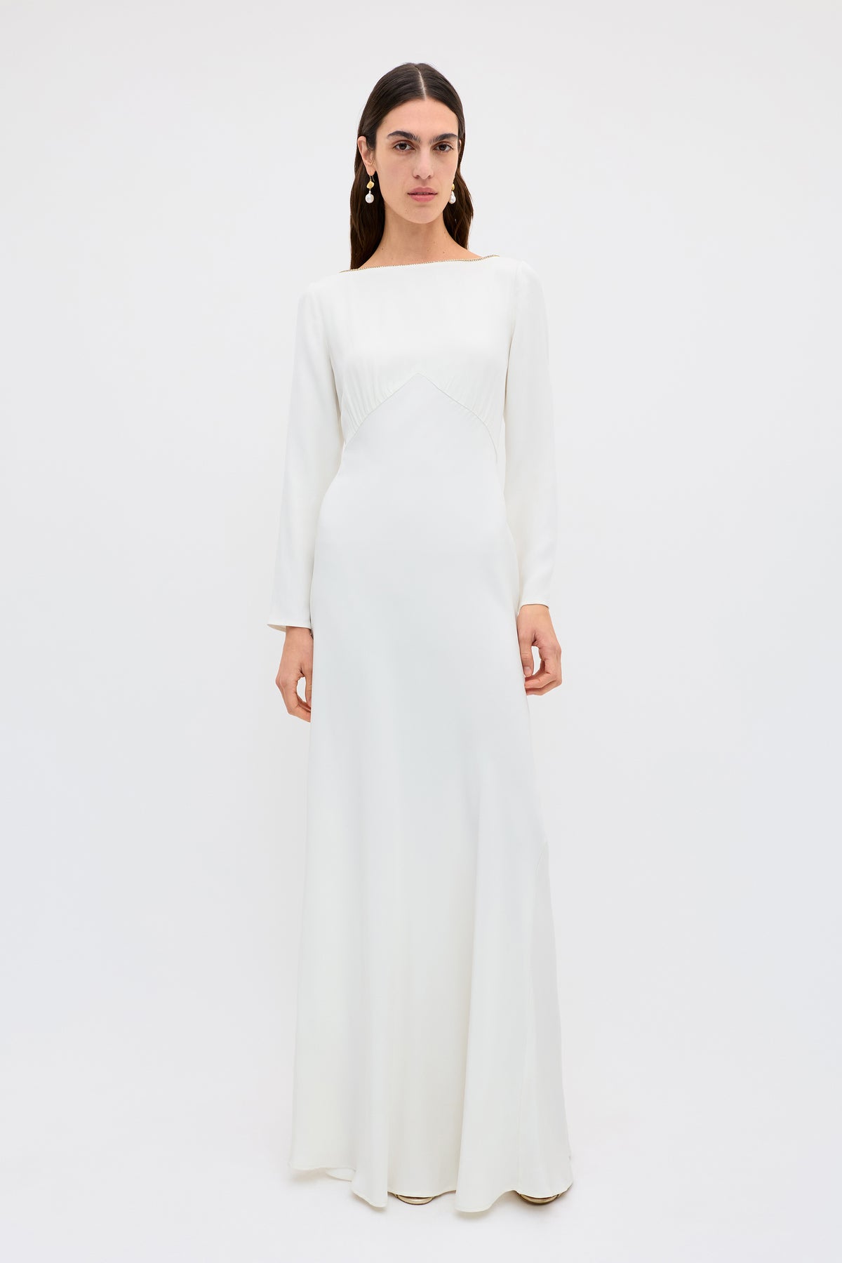A person with long, dark hair and drop earrings stands against a white background, wearing the Cesca - Ivory: a vintage-inspired, floor-length white silk gown featuring a fitted bodice and elegant boat neckline.