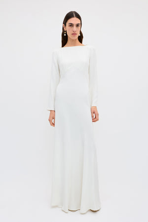 A person with long, dark hair and drop earrings stands against a white background, wearing the Cesca - Ivory: a vintage-inspired, floor-length white silk gown featuring a fitted bodice and elegant boat neckline.