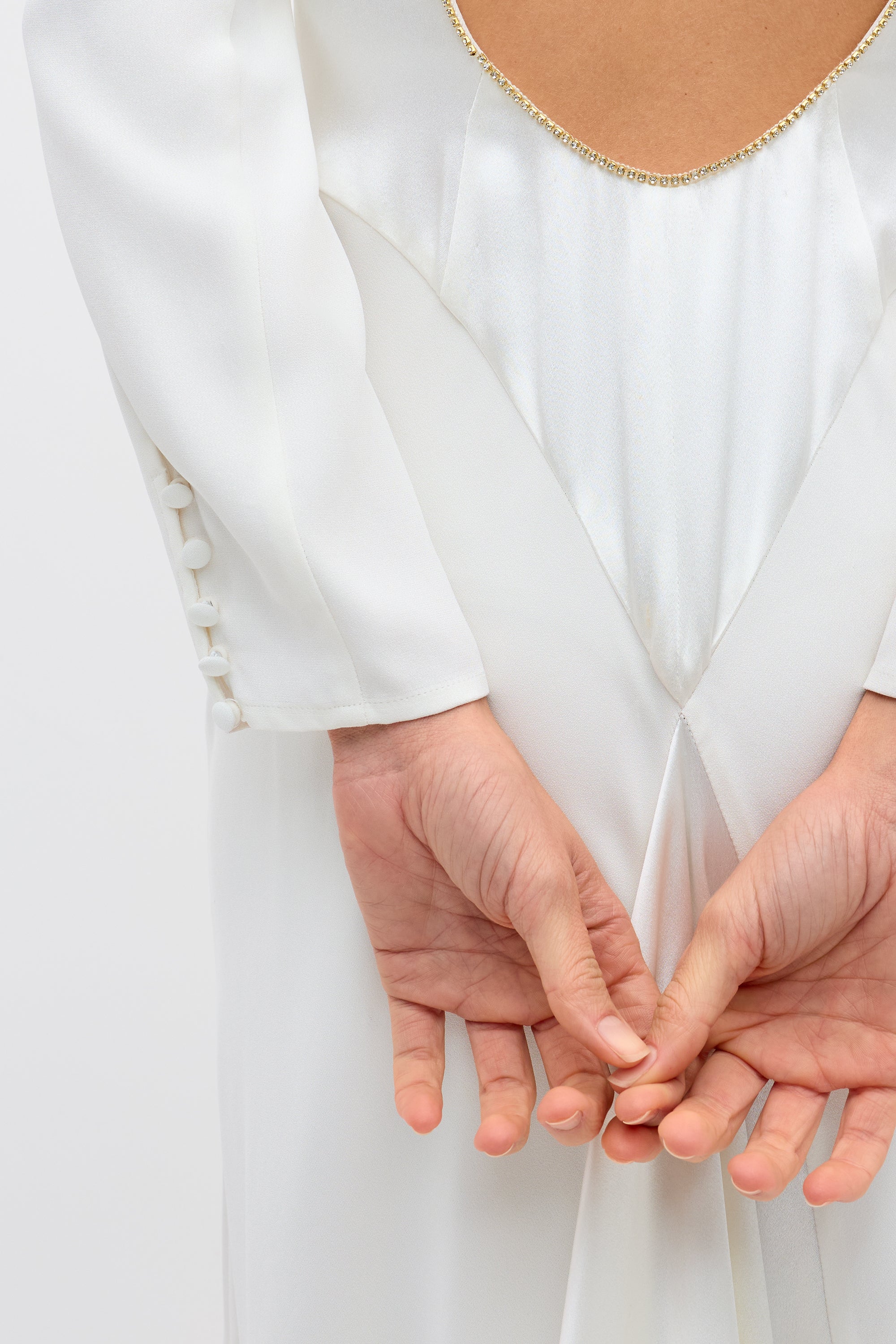A person wears the Cesca - Ivory: a vintage-inspired white silk gown with long sleeves, fabric-covered sleeve buttons, a deep gold-trimmed back, and a flowing satin train, standing with hands clasped behind their back.