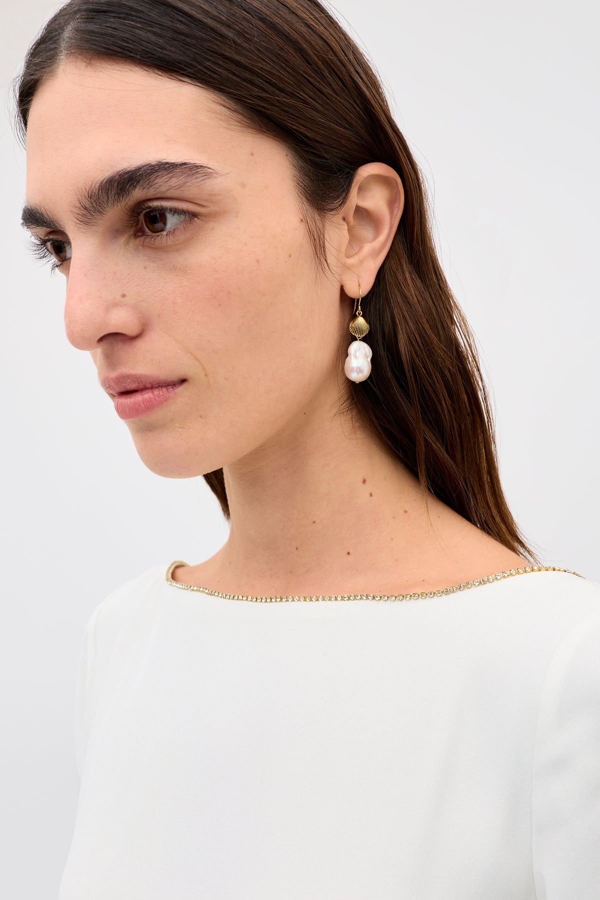 A woman with long dark hair wears the Cesca - Ivory top with a beaded neckline and a gold earring with a large white pearl, exuding vintage-inspired elegance as she gazes to the side against a plain light background.