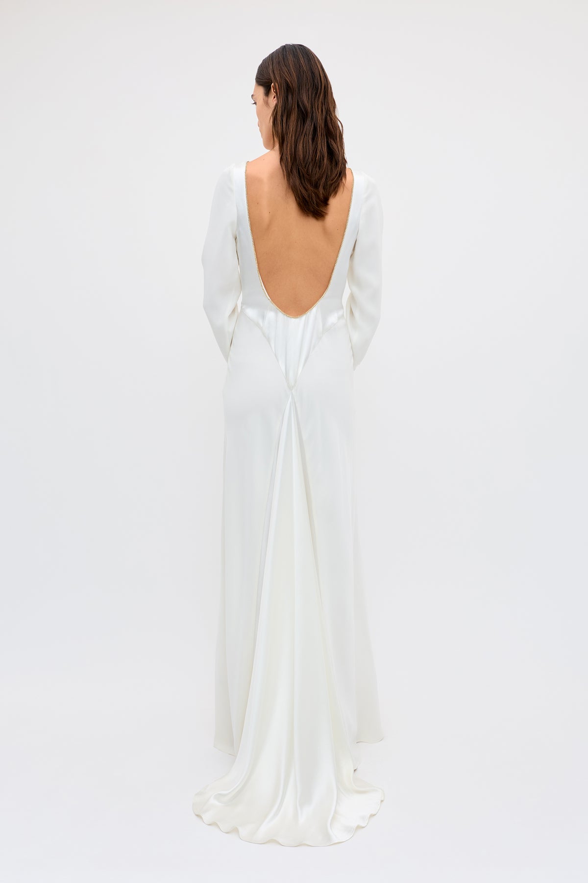 A woman with shoulder-length brown hair models the Cesca - Ivory: a vintage-inspired, long white satin dress with long sleeves, a dramatic low-cut back, and a flowing train, standing against a plain white background.