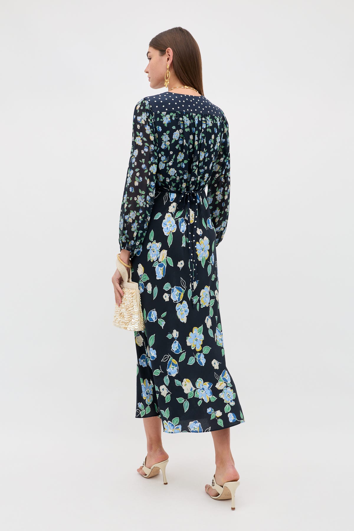 A woman faces away, wearing the Kaila - Layla Mix Navy long-sleeve, ankle-length black dress with blue and yellow floral patterns. She pairs it with a woven clutch, beige heeled sandals, and statement earrings.