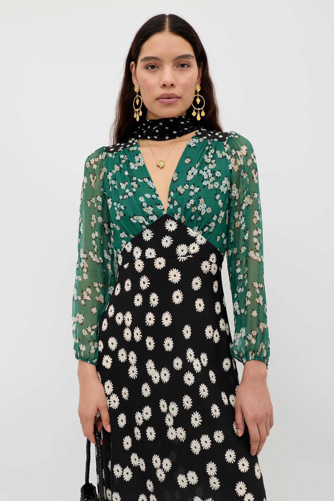 A woman with long dark hair models the Kaila - City Floral Mix Olive dress, showcasing bold green, black, and white floral prints. She accessorizes with gold earrings, a black choker with white dots, and a gold pendant necklace against a white background.