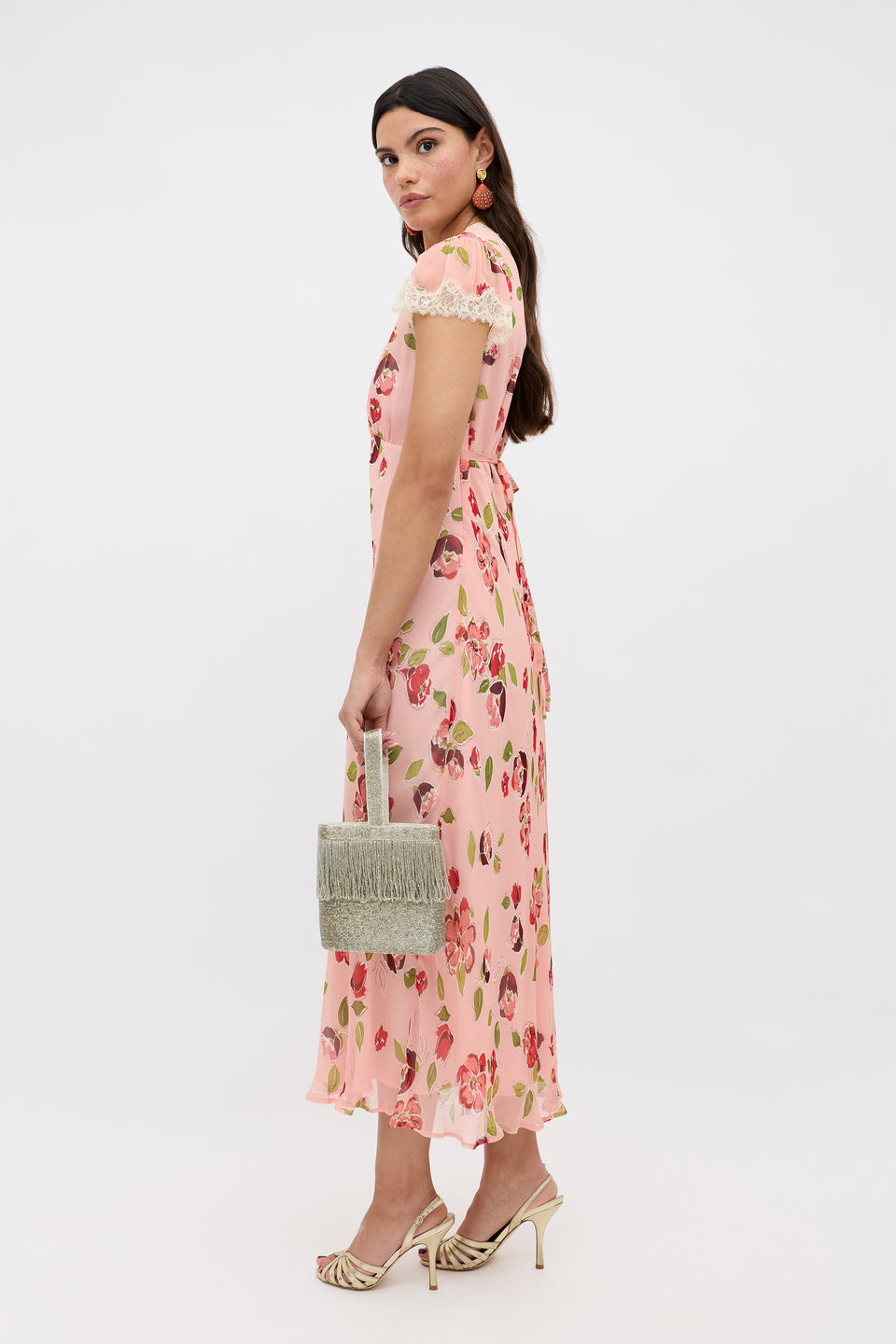 A woman in the Clarice - Kimmie Floral Pink midi dress with lace sleeves stands sideways, holding a small textured handbag. She wears beige heeled sandals, orange earrings, and has long dark hair over one shoulder against a plain background.