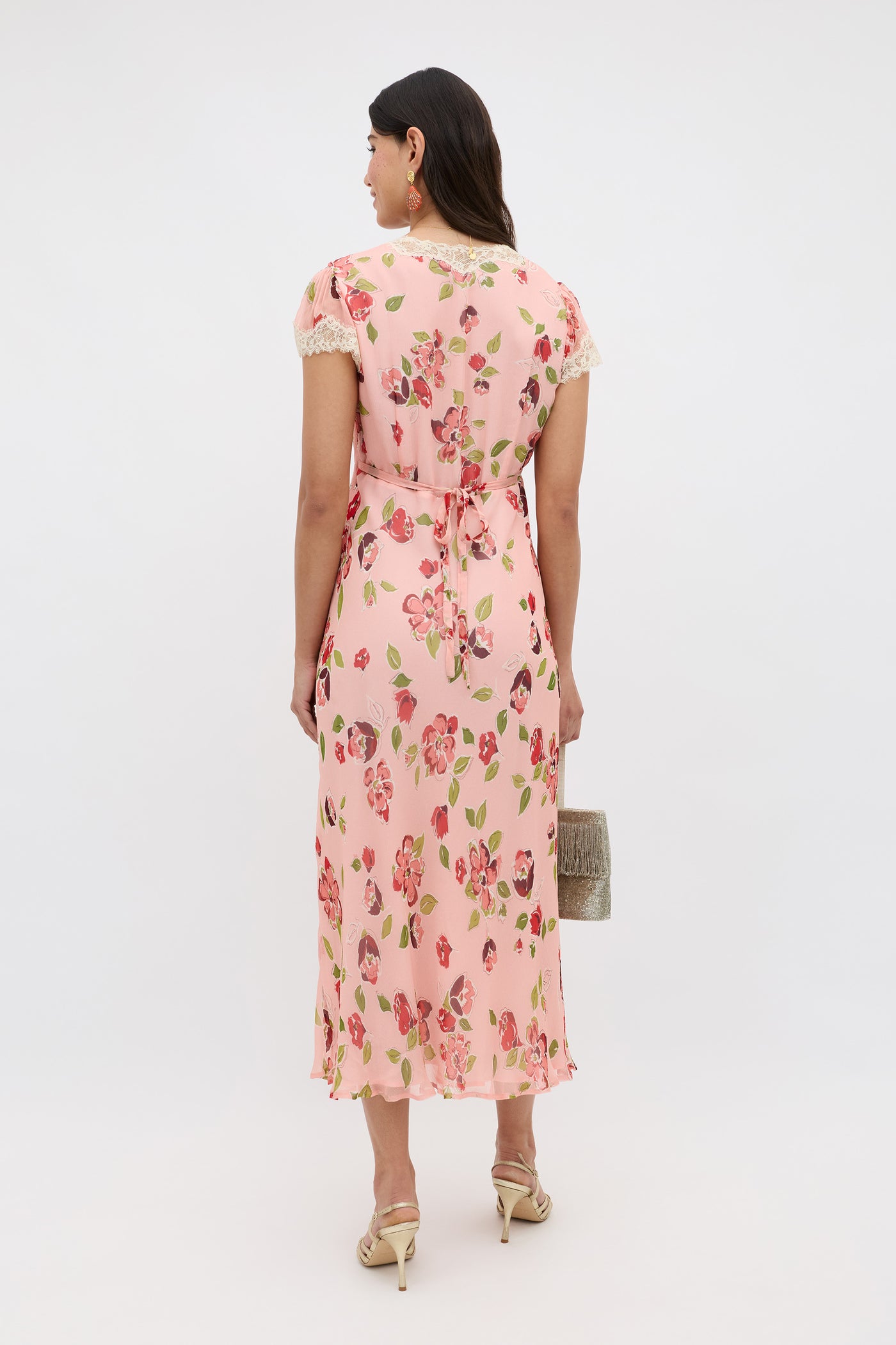 A woman faces away, wearing the Clarice - Kimmie Floral Pink midi dress with short sleeves and lace trim. She carries a small textured handbag, pairs beige high-heeled sandals, and her dark wavy hair is loose.