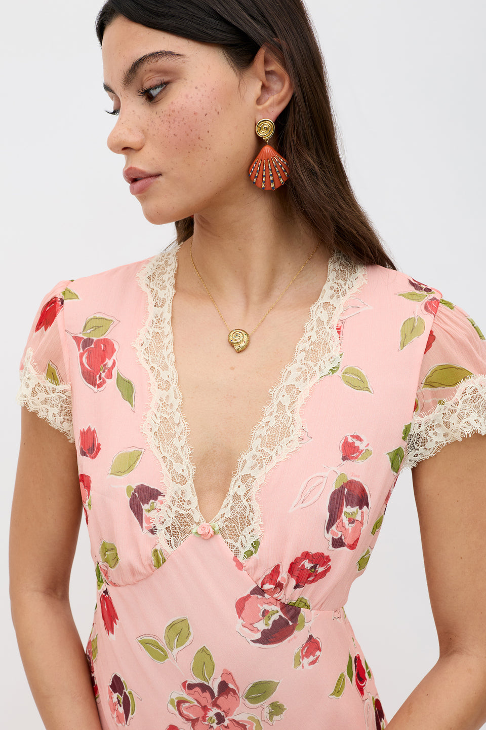 A woman with long brown hair wears the Clarice - Kimmie Floral Pink dress with lace trim, a gold necklace, and shell-shaped earrings. She gazes to the side against a plain light background.