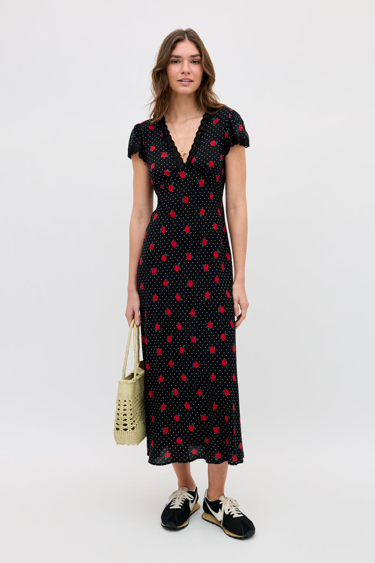 A woman stands against a plain background wearing the Clarice - Rose Spot Black midi dress with red polka dots, black sneakers, and holding a woven beige bag.