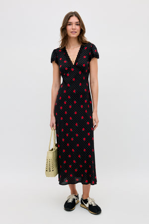 A woman stands against a plain background wearing the Clarice - Rose Spot Black midi dress with red polka dots, black sneakers, and holding a woven beige bag.